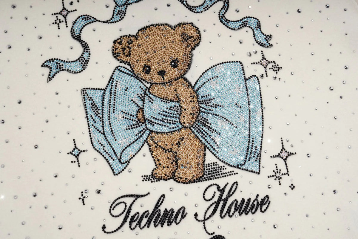 A close-up of a decorative surface featuring a teddy bear holding a large light blue bow, with small star motifs and the text 'Techno House' below on a white background with scattered rhinestone-like dots. TCH Rhinestone Teddy Bear Bow Graphic Cropped T-Shirt