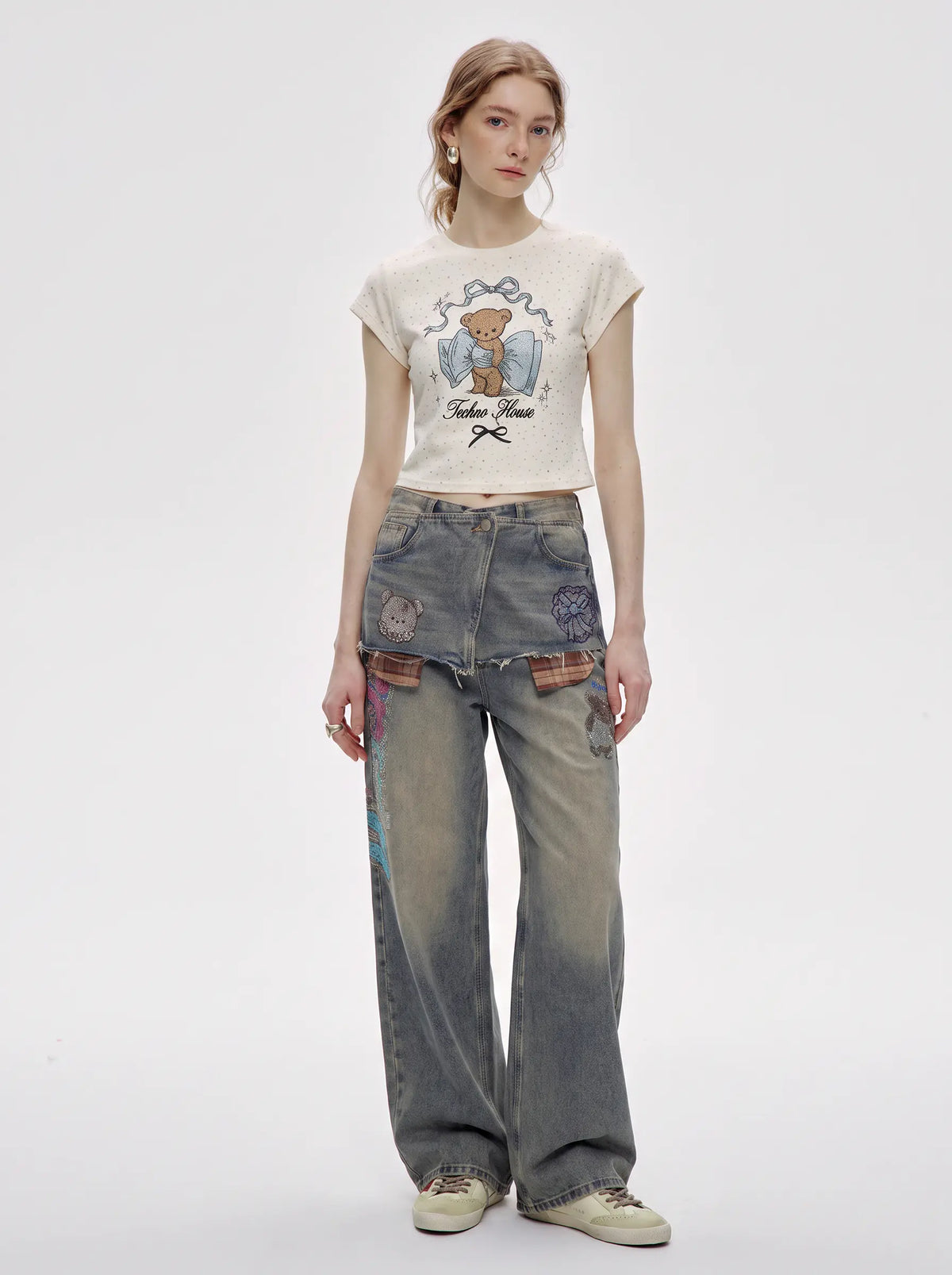 A model stands against a light gray studio background wearing a cream-colored cropped t-shirt with a teddy bear graphic and distressed, wide-leg gray jeans with colorful patch details on the pockets. TCH Rhinestone Teddy Bear Bow Graphic Cropped T-Shirt