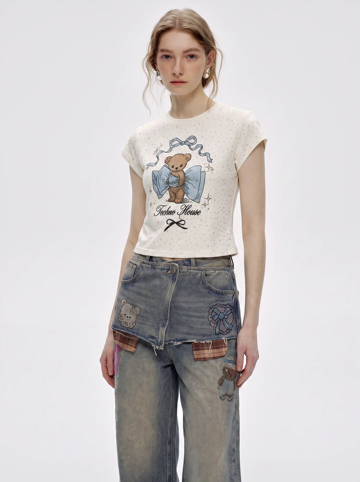 A model wearing a short-sleeve cream-colored t-shirt with a teddy bear graphic and 'Faken Heine' text, paired with distressed denim jeans, against a plain light gray background. TCH Rhinestone Teddy Bear Bow Graphic Cropped T-Shirt