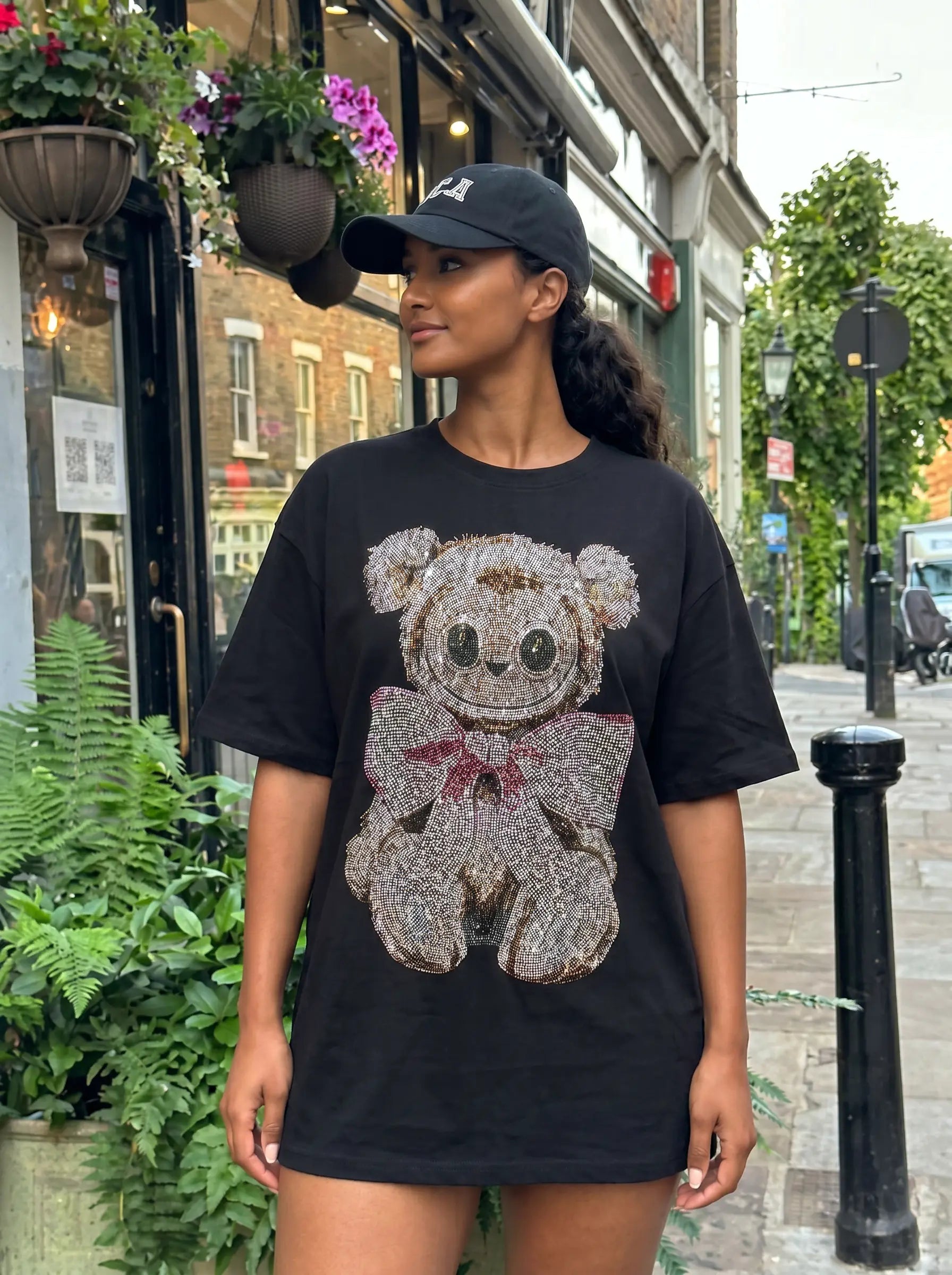A woman wearing a black oversized t-shirt with a teddy bear graphic made of rhinestones, standing on a city street. TCH Rhinestone Teddy Bear Bow Graphic Oversized Black T-Shirt