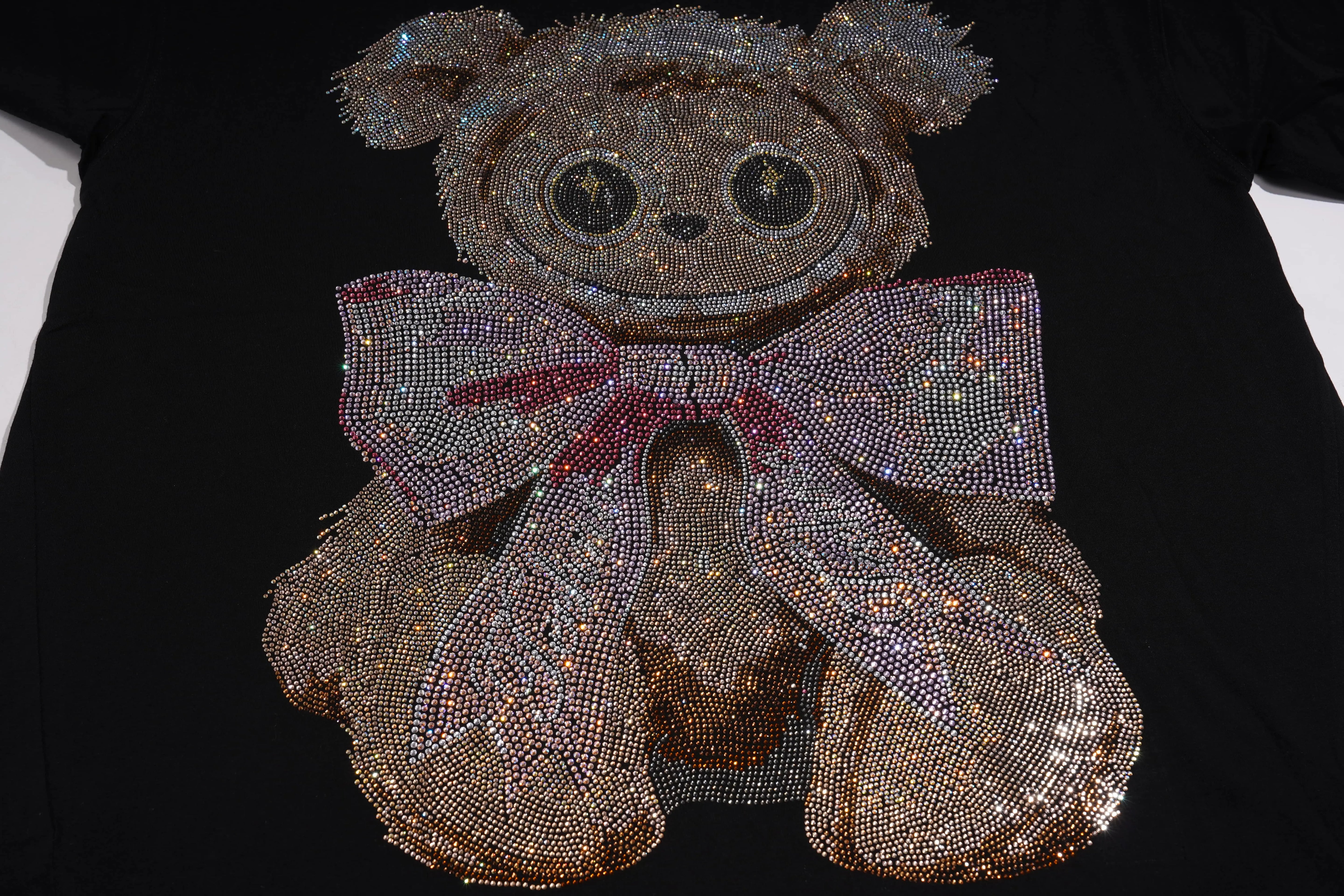 A black T-shirt featuring a large teddy bear design embellished with sparkling rhinestones, complete with a glittering pink and silver bow, shown on a white background. TCH Rhinestone Teddy Bear Bow Graphic Oversized Black T-Shirt