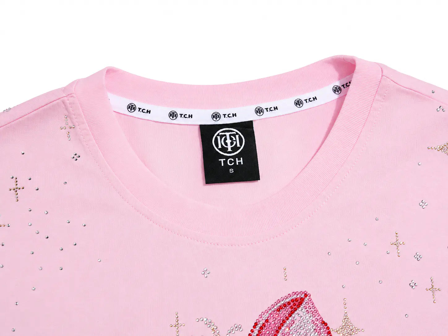 Close-up of a light pink crewneck sweatshirt featuring a black TCH neck label and a white inner collar band with repeating TCH logos. The fabric is decorated with small silver rhinestones and gold cross motifs. TCH Rhinestone Teddy Bear Bow Pink Graphic T-Shirt
