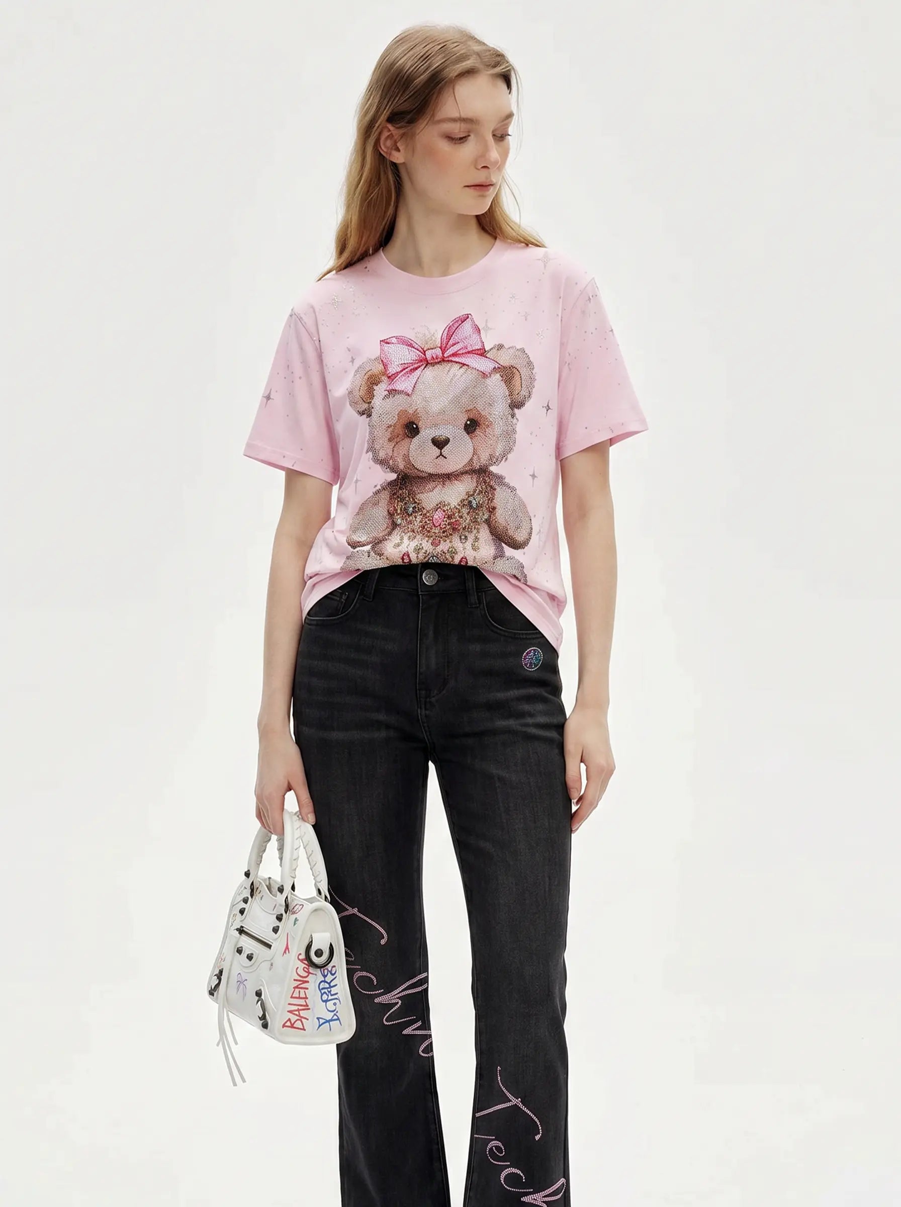 A model wearing a light pink graphic t-shirt with a teddy bear print, paired with black jeans and holding a white patterned handbag against a plain white background. TCH Rhinestone Teddy Bear Bow Pink Graphic T-Shirt