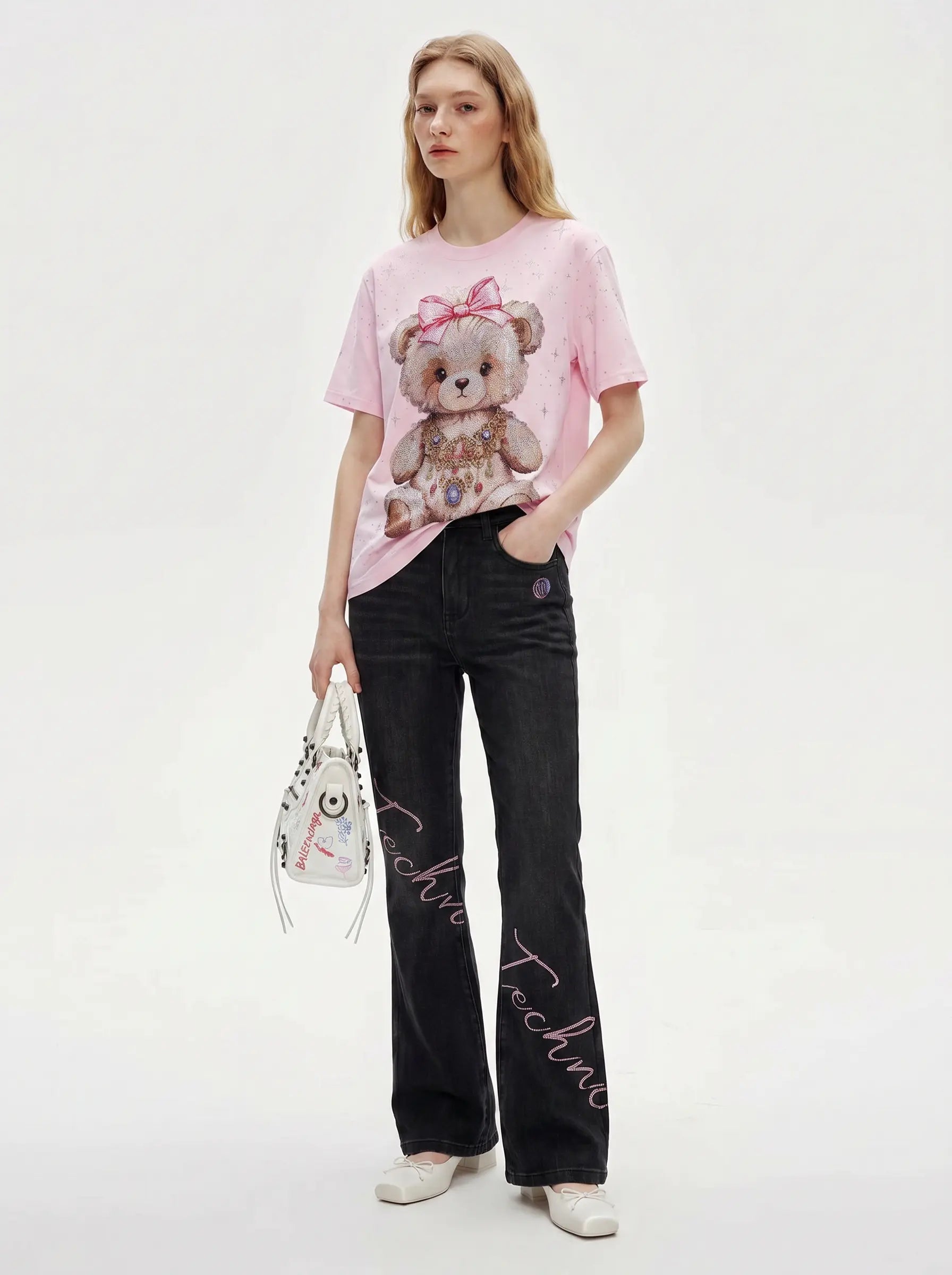 A model stands against a light gray studio background wearing a light pink graphic T-shirt with a teddy bear print, black flared jeans with script embroidery down the legs, and holding a white handbag. TCH Rhinestone Teddy Bear Bow Pink Graphic T-Shirt