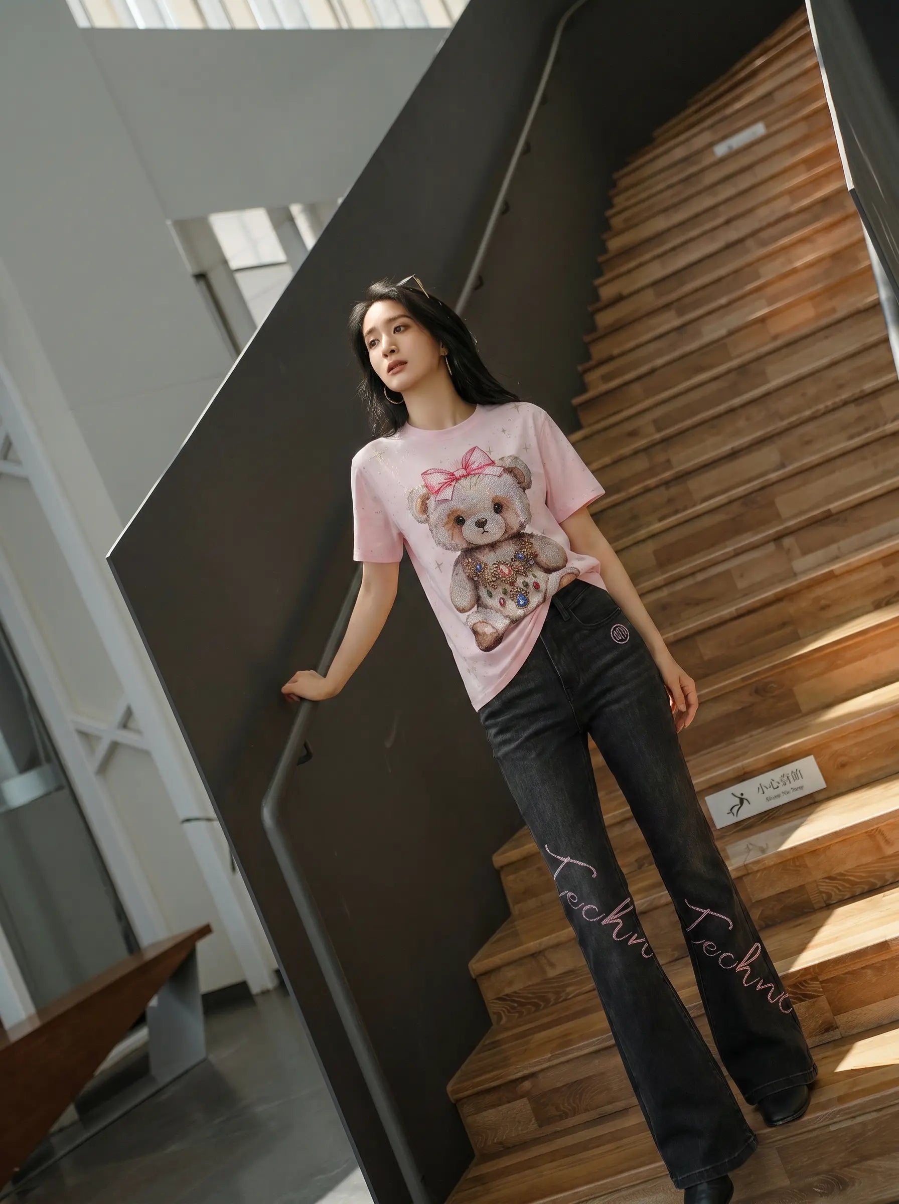A woman stands on an indoor wooden staircase wearing a light pink graphic T-shirt with a teddy bear print and dark flare jeans. TCH Rhinestone Teddy Bear Bow Pink Graphic T-Shirt
