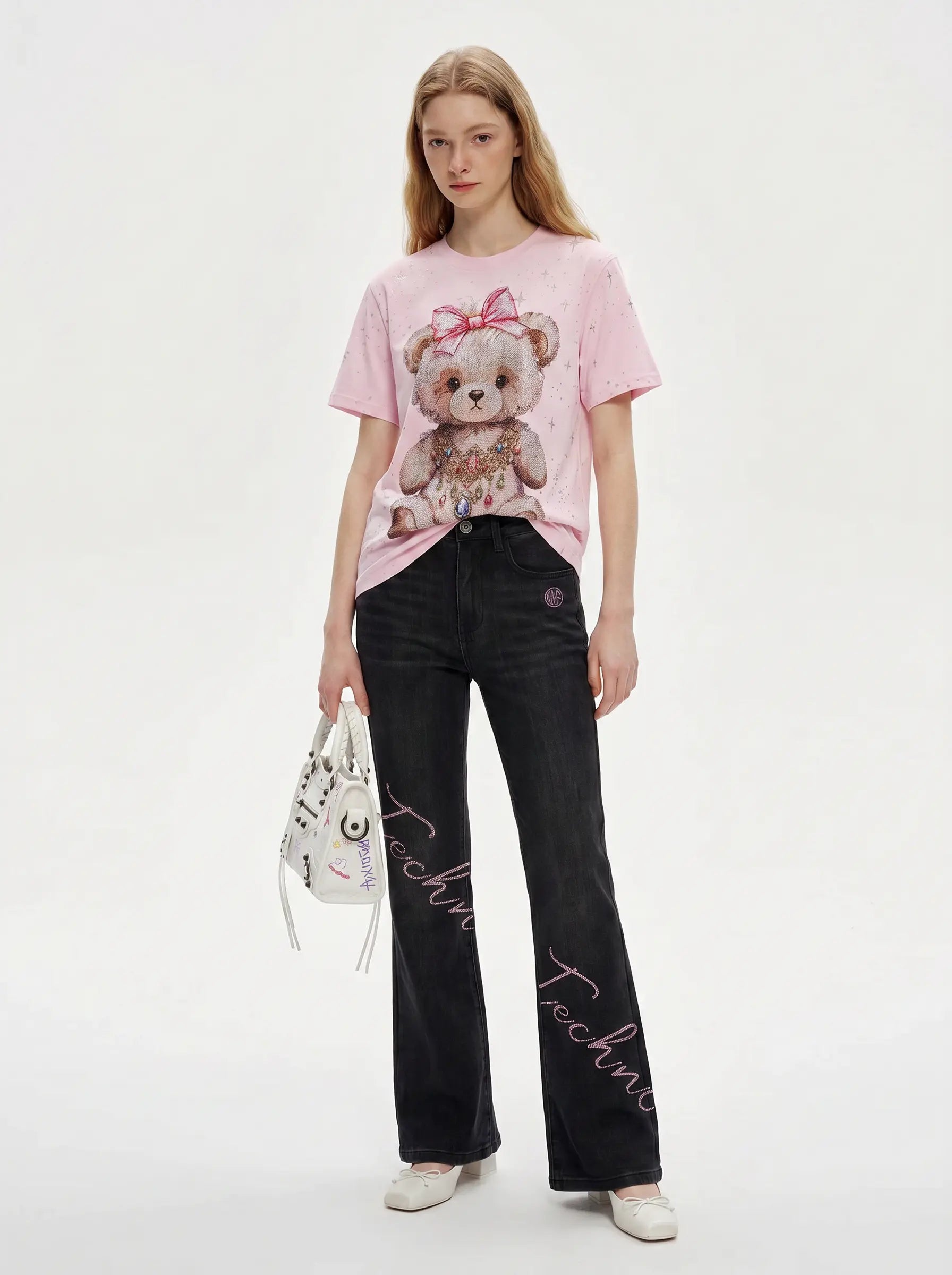 A model stands against a light gray studio background wearing a light pink graphic T-shirt with a teddy bear print, black wide-leg jeans with white script text down the legs, and white shoes. She is holding a small white handbag. TCH Rhinestone Teddy Bear Bow Pink Graphic T-Shirt