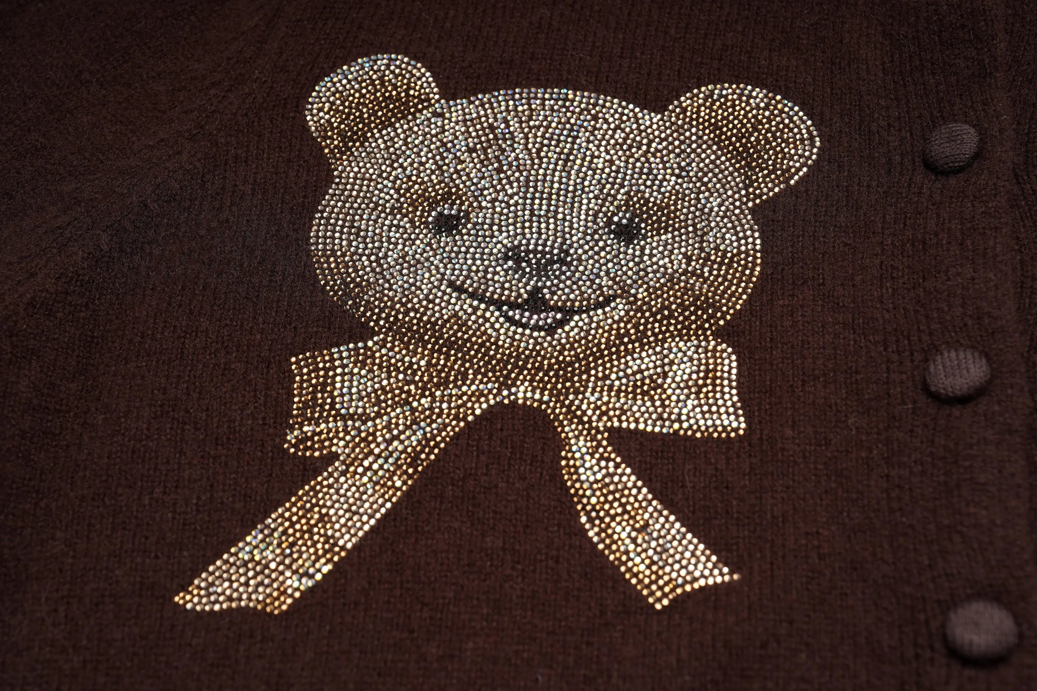 A close-up of a dark brown knit garment featuring a teddy bear face and bow embellished with shimmering gold rhinestones, with three matching gold buttons visible on the right side. TCH Rhinestone Teddy Bear Bow Ruffle Knit Cardigan