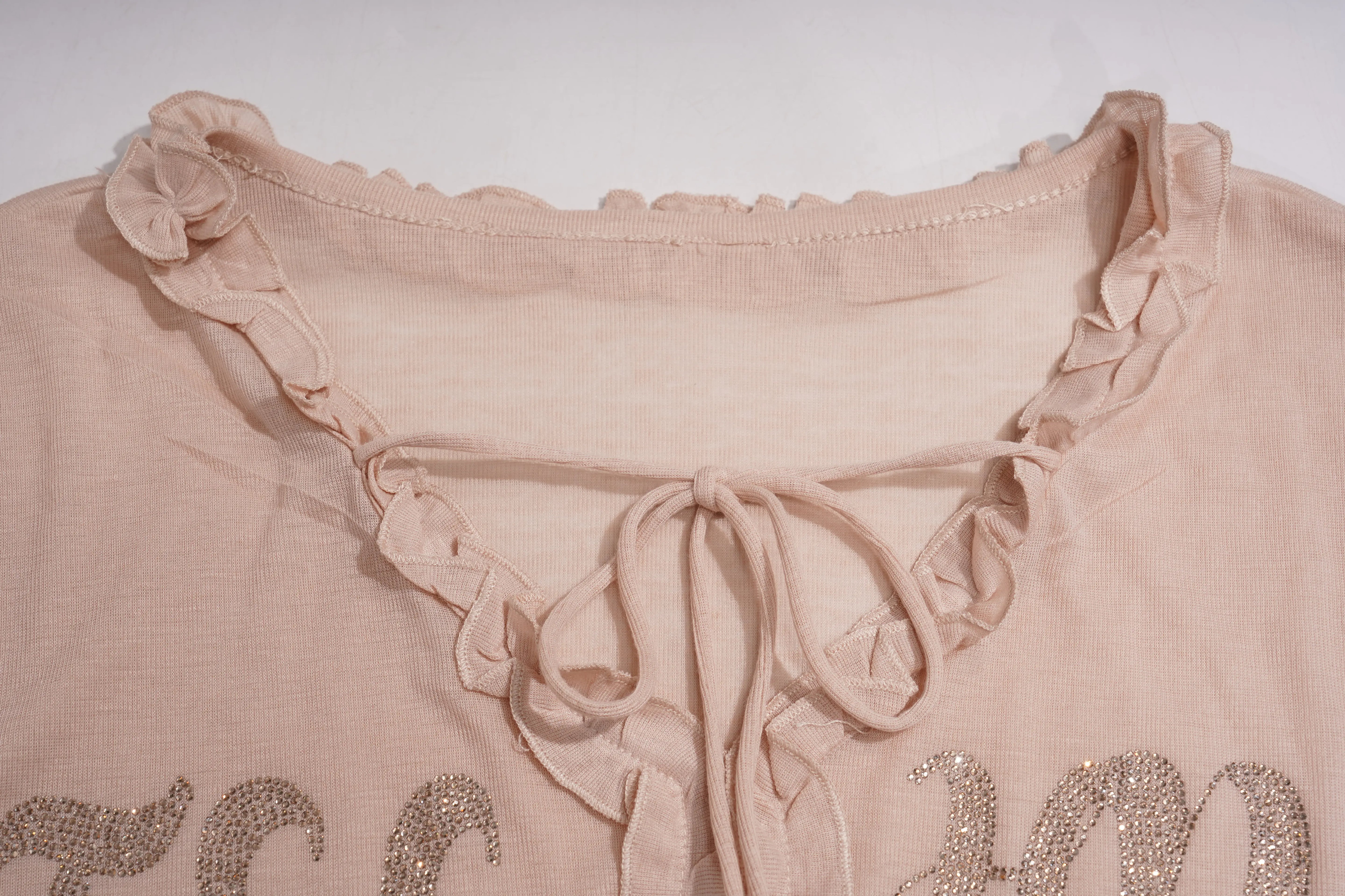 Close-up of a light beige or blush pink blouse with a ruffled lace-up neckline and sequined lettering at the bottom. TCH Rhinestone Teddy Bear Bow Ruffle Long Sleeve Top