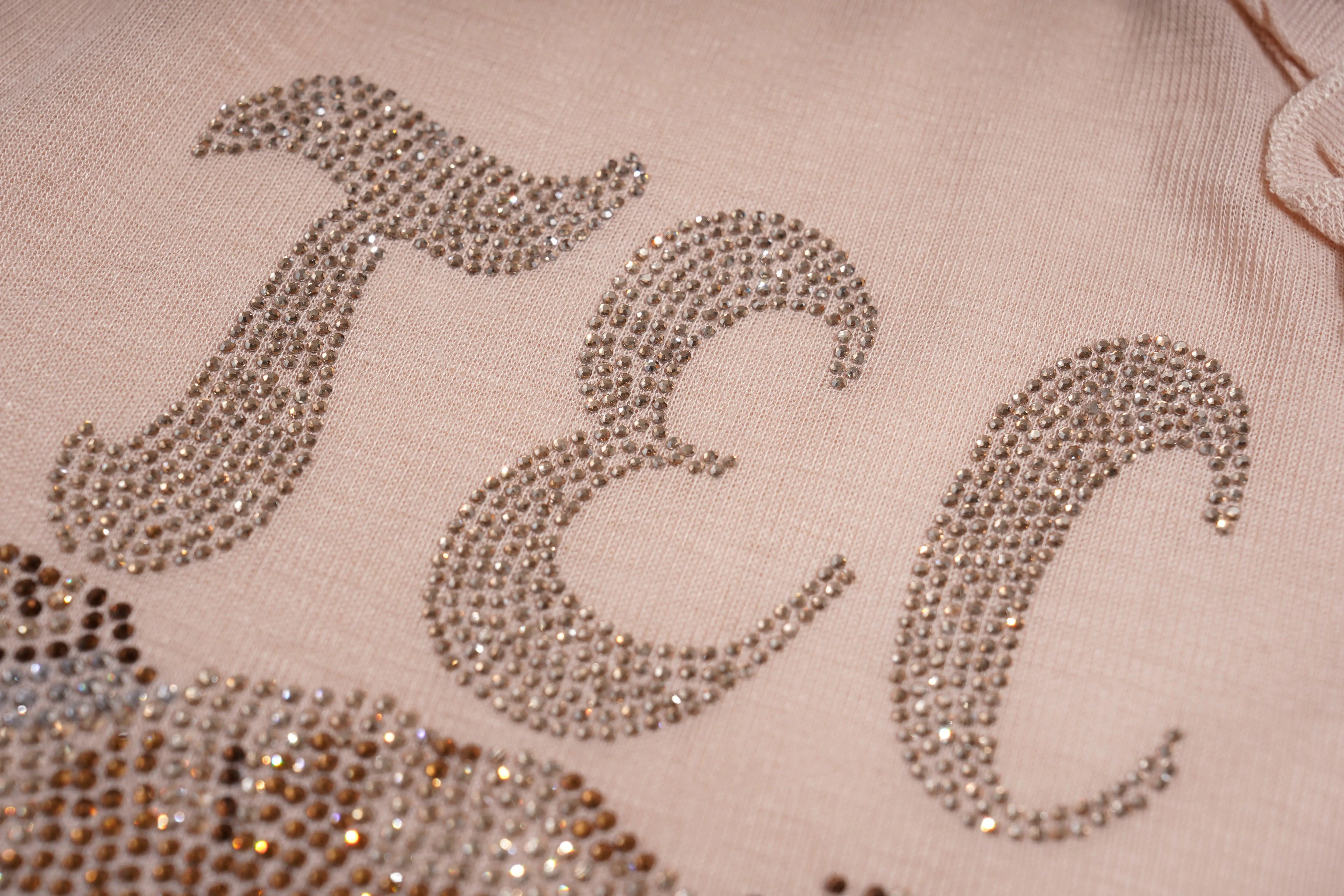 Close-up of a light pink fabric with large silver rhinestone letters 'JEC' and part of a matching rhinestone design at the bottom left. TCH Rhinestone Teddy Bear Bow Ruffle Long Sleeve Top
