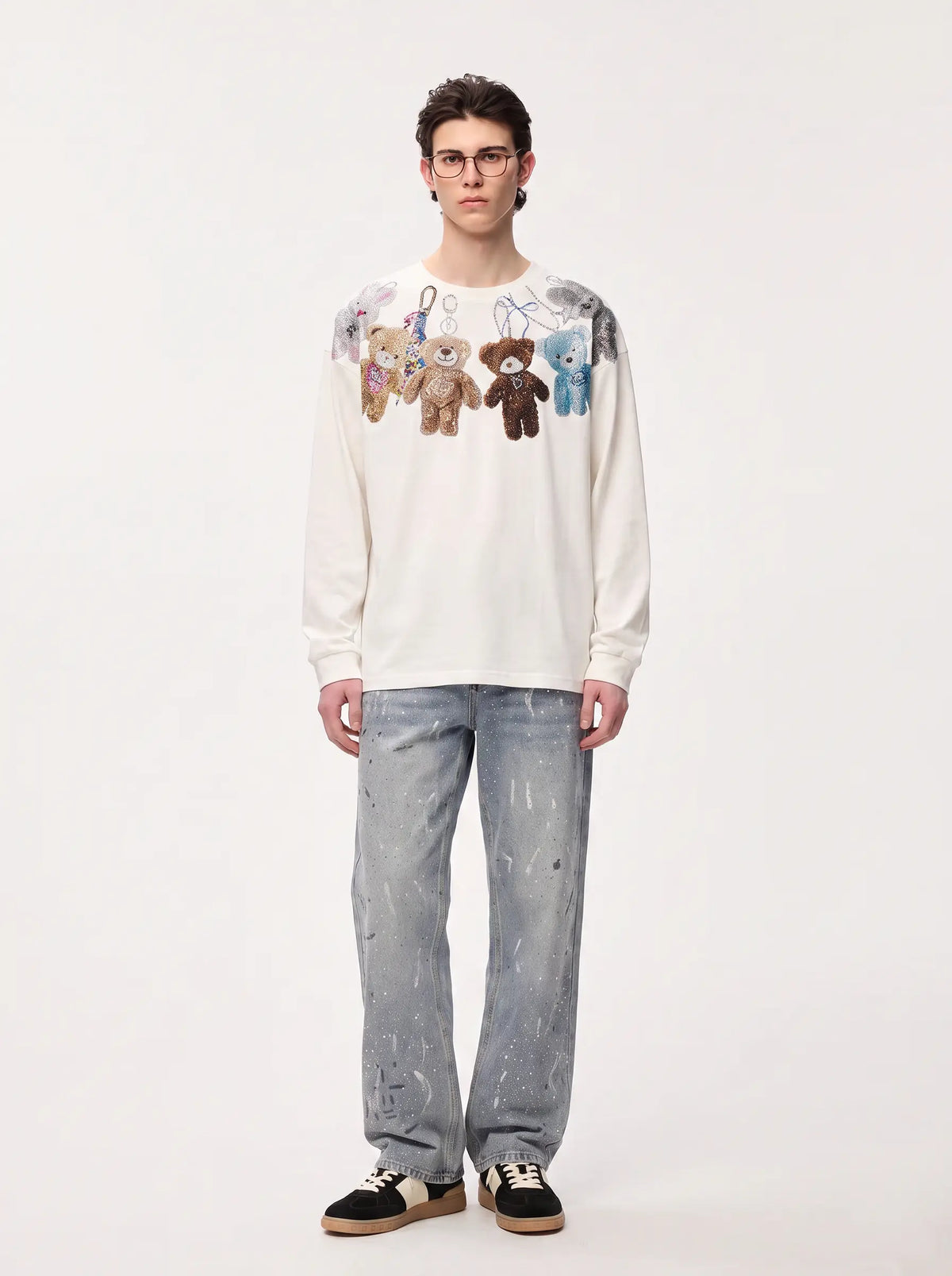A model stands against a plain light background wearing a cream-colored long-sleeve sweatshirt with a graphic of hanging teddy bears on the chest, paired with light-wash jeans and black-and-white sneakers. TCH Rhinestone Teddy Bear Charm Graphic Long Sleeve Sweatshirt