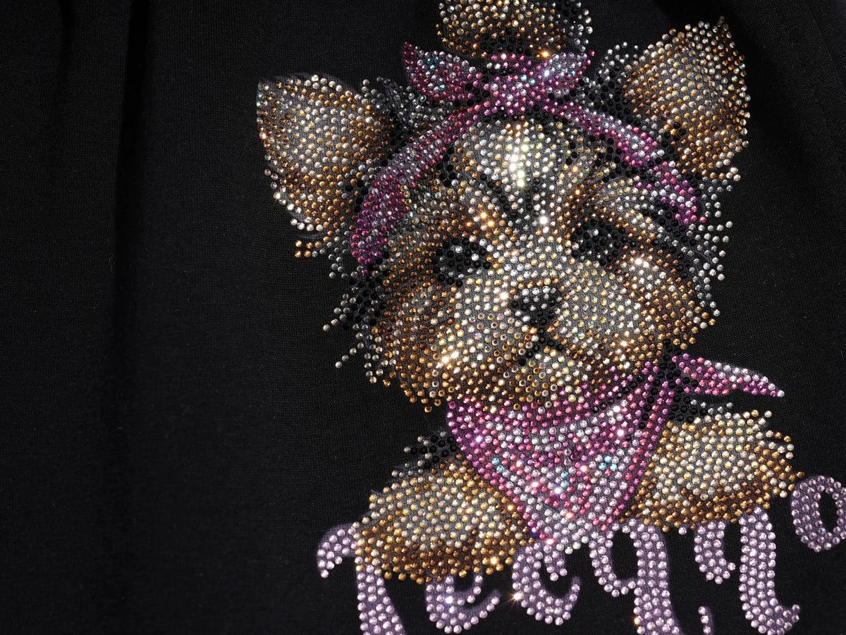 A close-up of a black garment featuring a large, highly embellished graphic of a puppy made of gold, silver, and pink rhinestones, with the word 'petite' written below in matching pink and silver rhinestones. TCH Rhinestone Teddy Bear Cherry Graphic Sweat Shorts