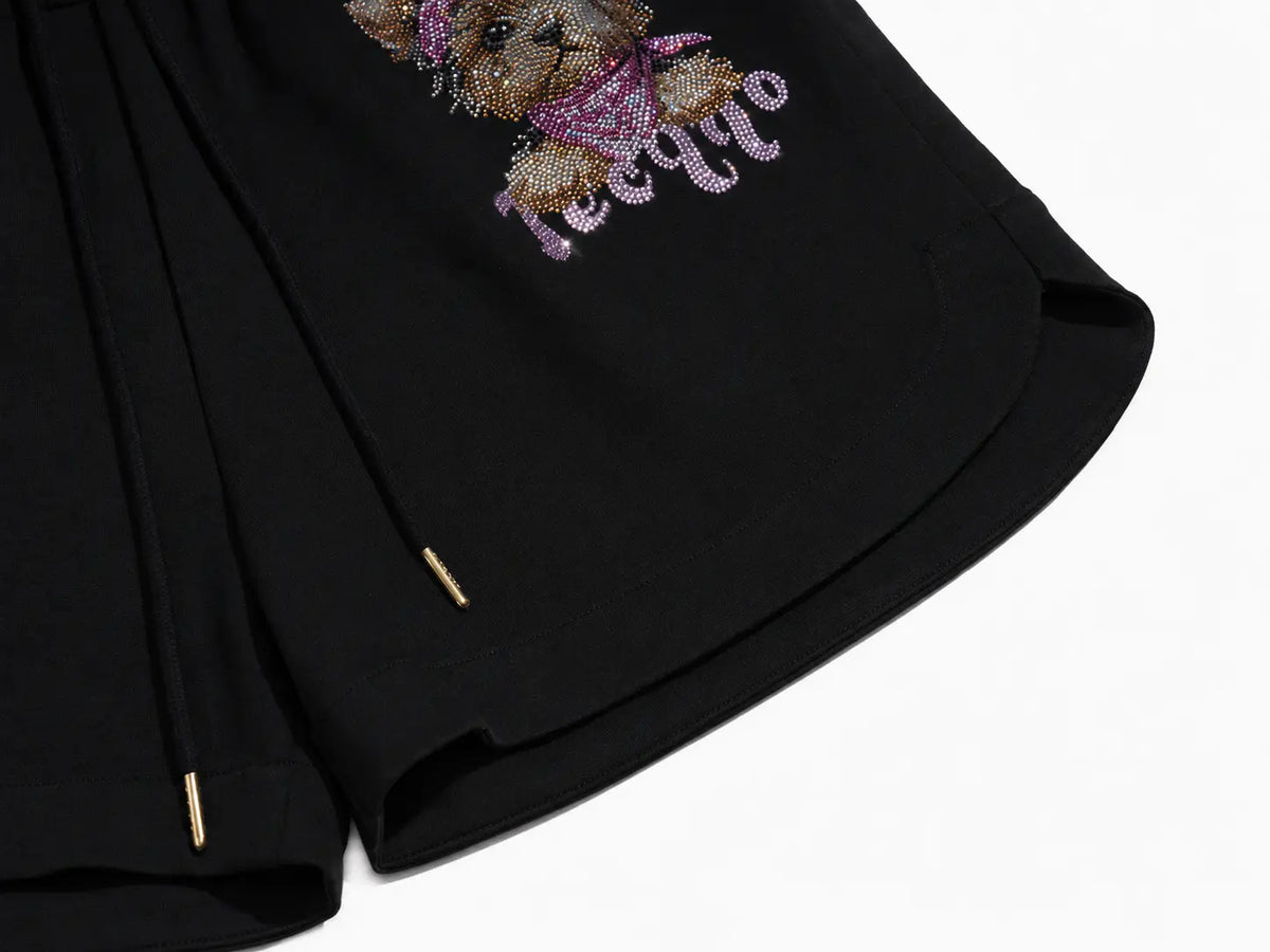 Close-up of black shorts with a decorative embroidered teddy bear design in metallic gold and purple rhinestones on the upper left leg, plus small gold zipper pulls. TCH Rhinestone Teddy Bear Cherry Graphic Sweat Shorts