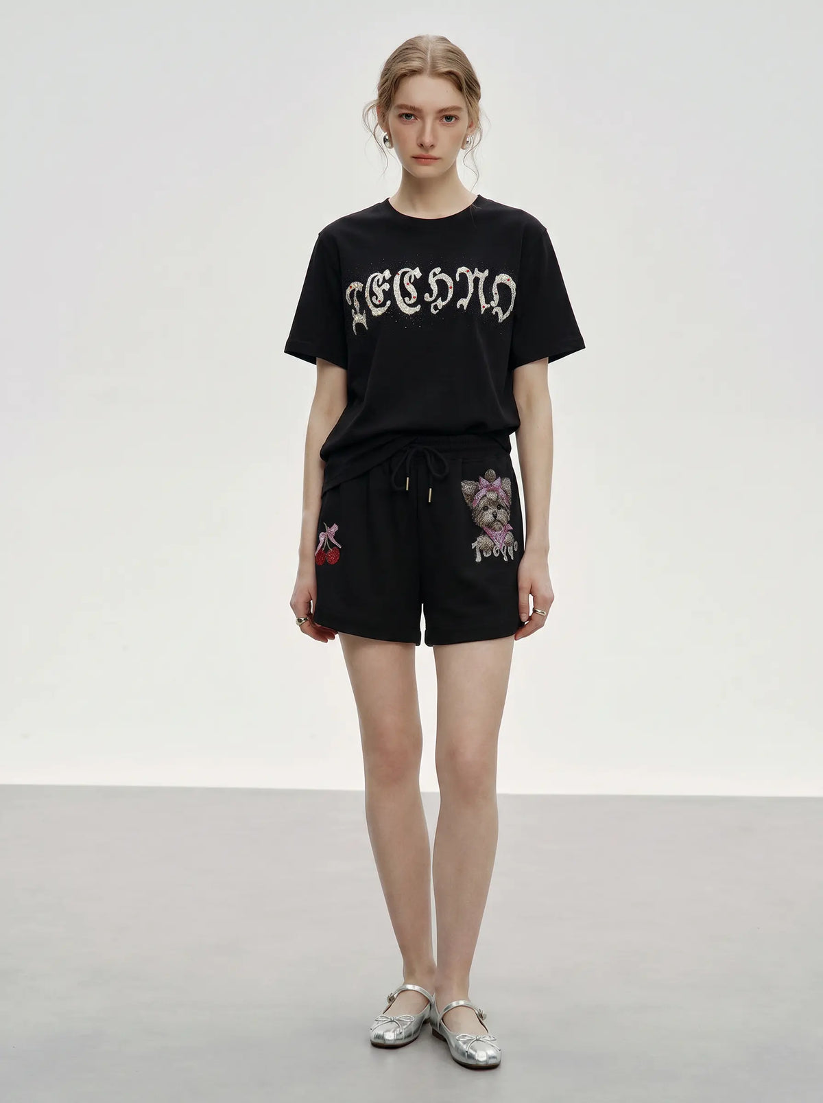 A model stands against a light gray studio background wearing a black graphic T-shirt with white gothic lettering and black shorts with colorful floral embroidery on the sides. She is wearing silver flat shoes. TCH Rhinestone Teddy Bear Cherry Graphic Sweat Shorts