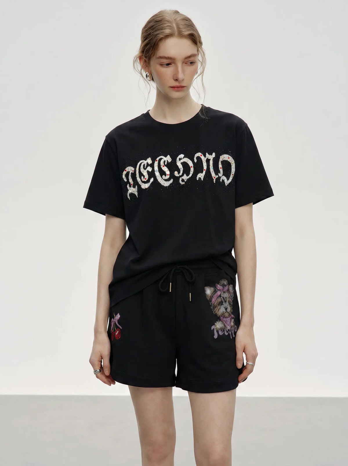 A model wearing a black short-sleeve t-shirt with a white gothic-style graphic print and black shorts featuring floral embroidery, set against a plain light gray studio background. TCH Rhinestone Teddy Bear Cherry Graphic Sweat Shorts