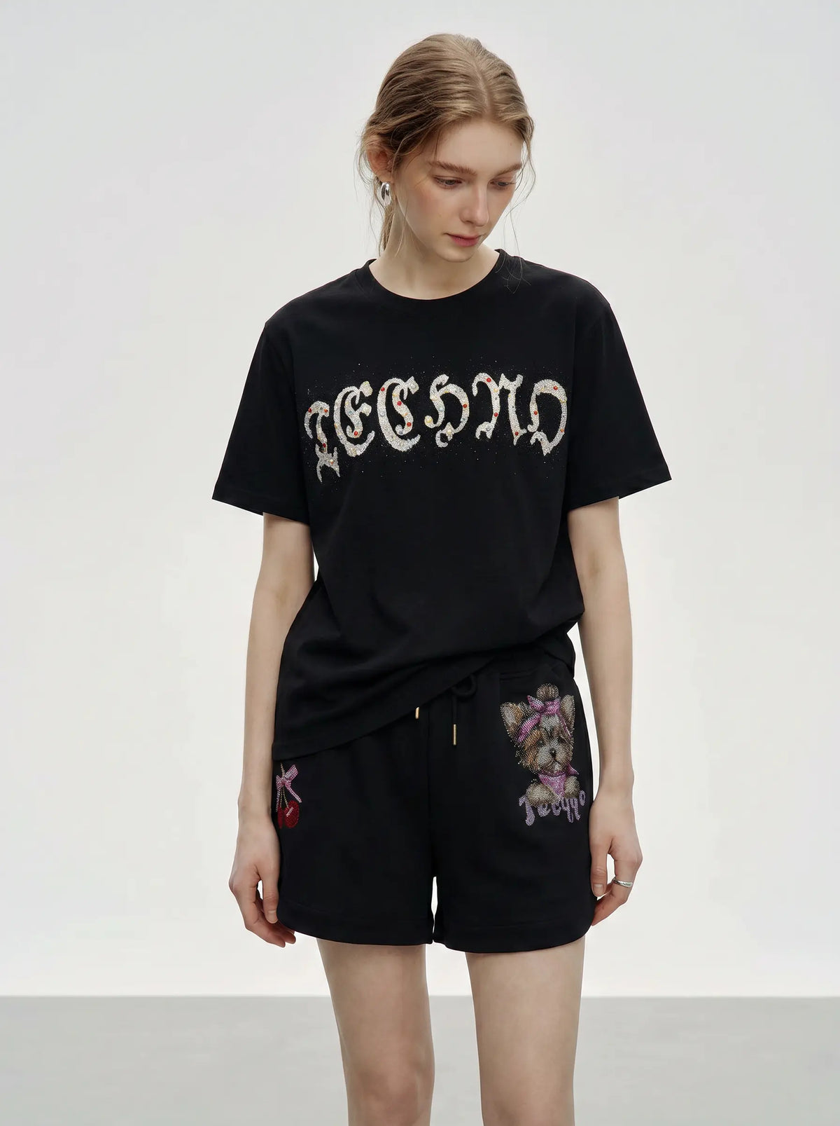 A model wearing a black short-sleeve t-shirt with a gothic-style white graphic print and black shorts featuring purple floral embroidery, set against a plain light gray studio background. TCH Rhinestone Teddy Bear Cherry Graphic Sweat Shorts