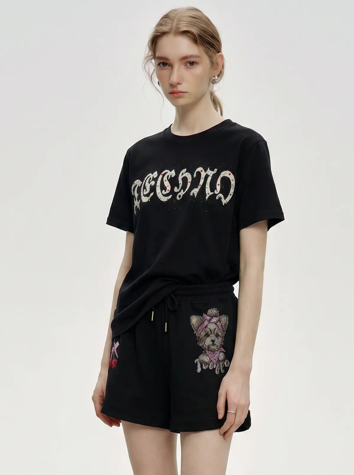 A model wearing a black short-sleeve t-shirt with a gothic-style embroidered graphic and black shorts featuring a dog patch, set against a plain light gray background. TCH Rhinestone Teddy Bear Cherry Graphic Sweat Shorts