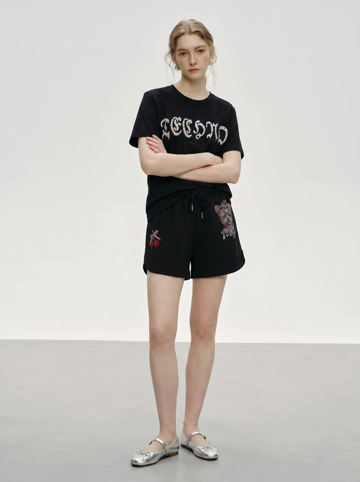 A model stands in a studio wearing a black short-sleeve t-shirt with 'TECHNO' in white gothic lettering, paired with matching black shorts featuring cherry embroidery, and silver flats. TCH Rhinestone Teddy Bear Cherry Graphic Sweat Shorts