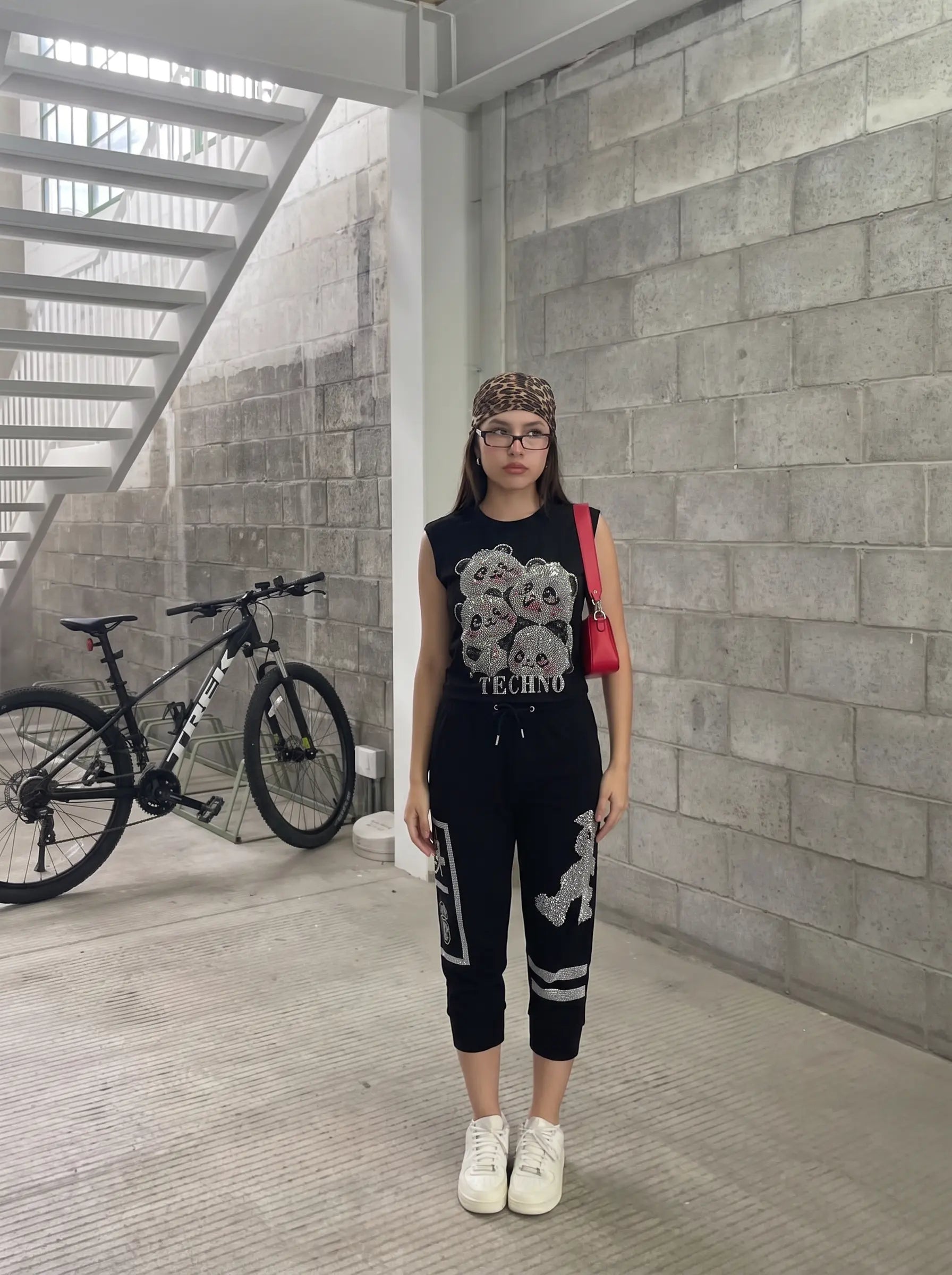 A woman stands in a concrete stairwell wearing a black sleeveless graphic top with a skull print and the word 'TECHNO' on the front, paired with black jogger pants featuring white graphic prints, white sneakers, and a red shoulder bag. TCH Rhinestone Teddy Bear Graphic Black Jogger Pants