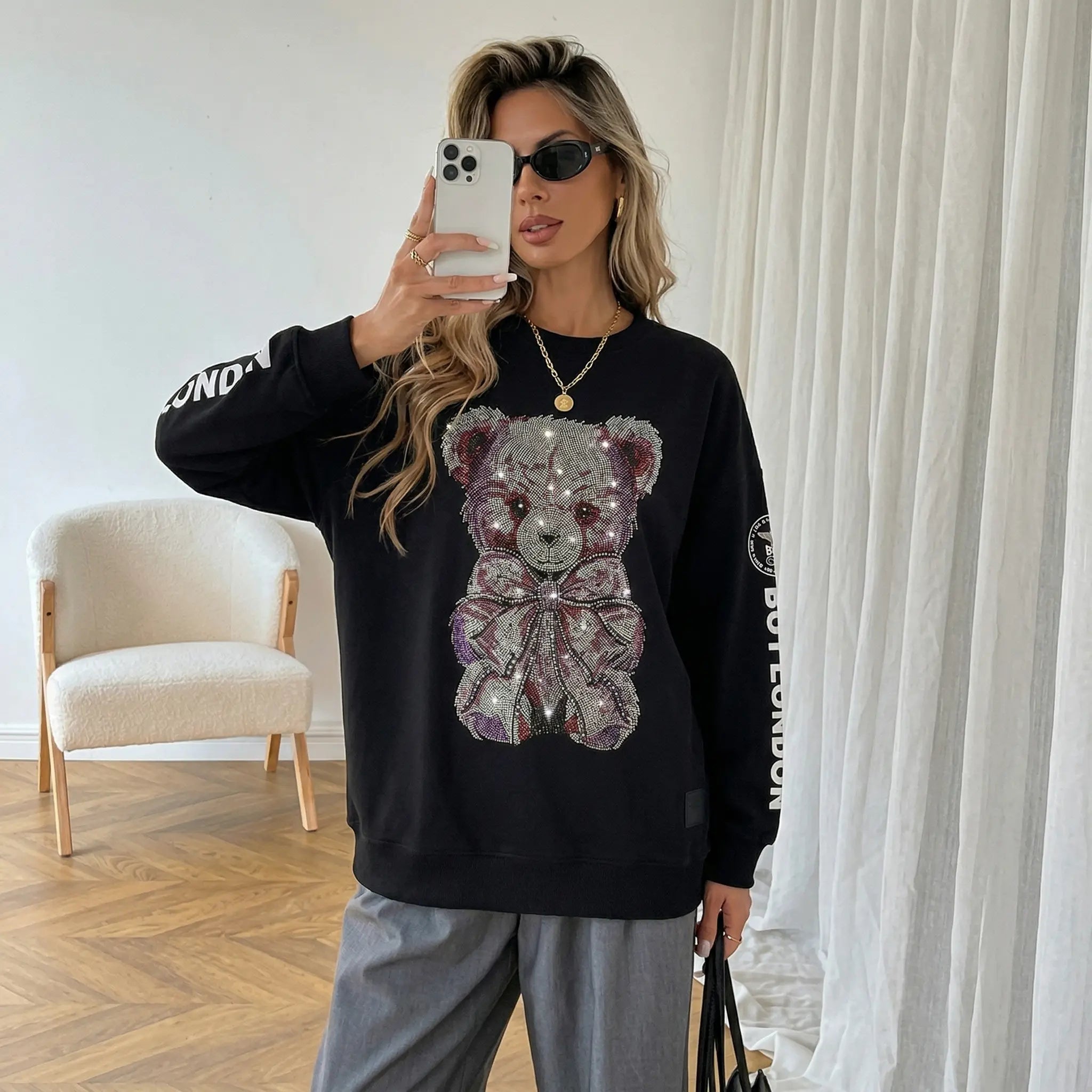 Woman wearing a black sweatshirt with a teddy bear design, taking a mirror selfie in a room.  TCH Rhinestone Teddy Bear Graphic Crewneck Sweatshirt-
