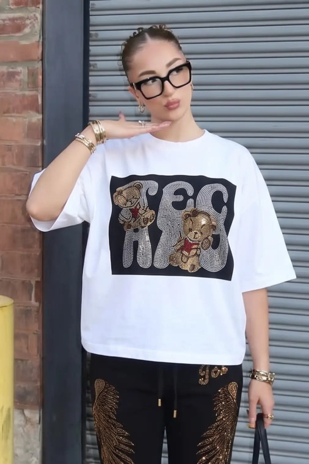 A woman stands outdoors in front of a gray metal garage door and brick wall, wearing an oversized white graphic T-shirt with a black chest print, black pants with gold wing and stripe graphics, black sneakers, and carrying a small black handbag. TCH Rhinestone Teddy Bear Graphic White T-Shirt