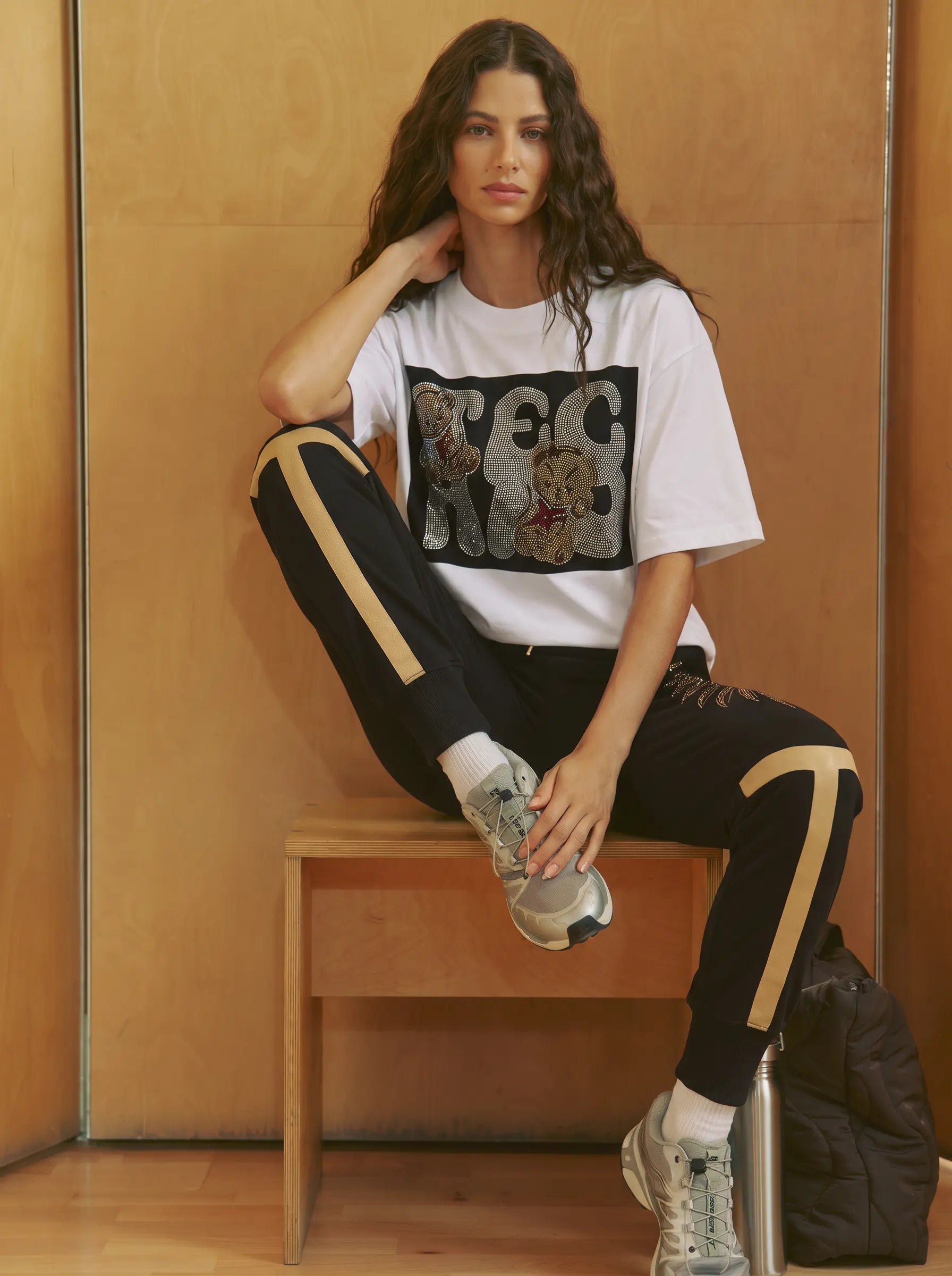 A woman sits on a wooden bench wearing a white graphic t-shirt with a black and gray bear print, black track pants with beige side stripes, and gray sneakers, set against a warm wood-paneled background. TCH Rhinestone Teddy Bear Graphic White T-Shirt