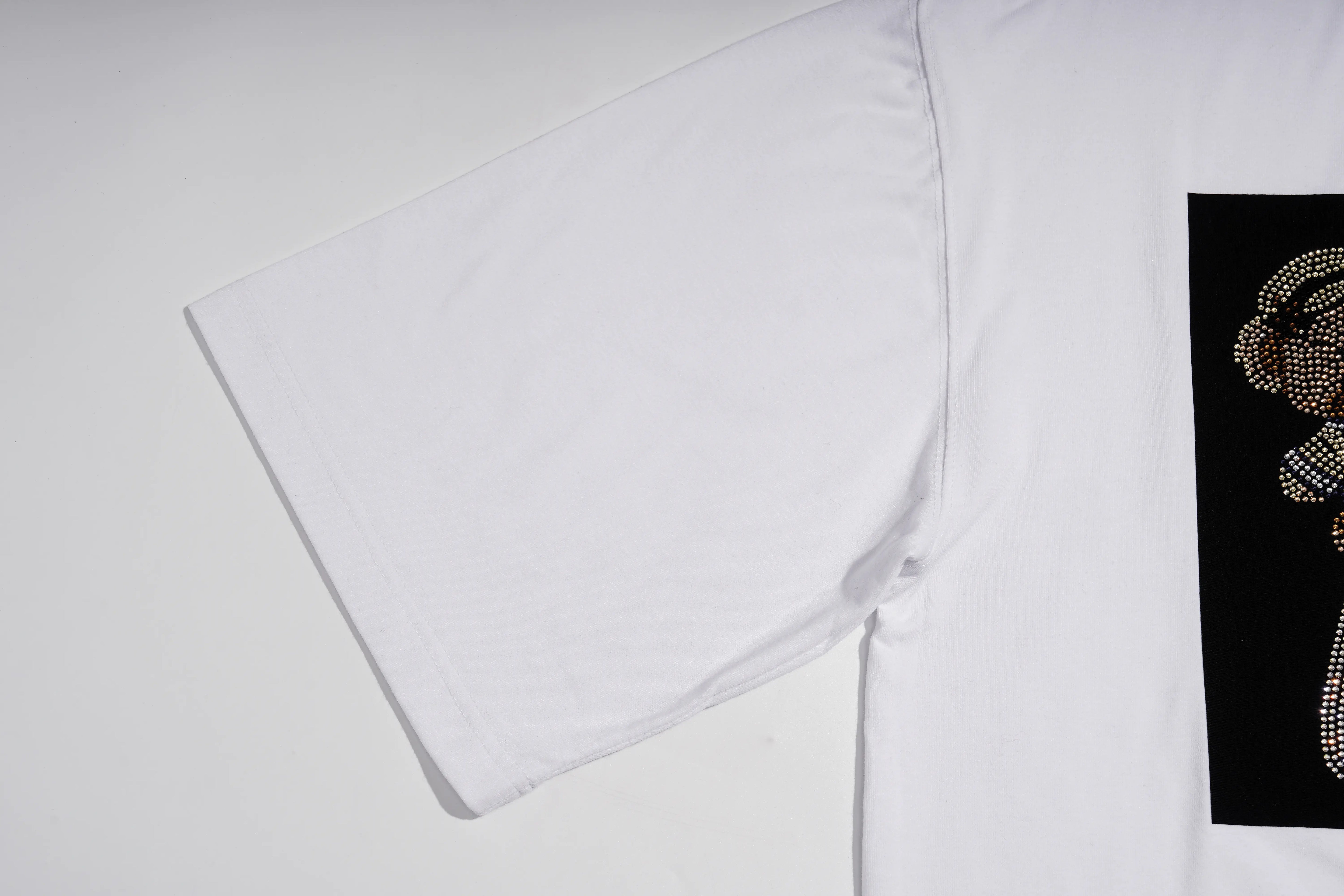 A close-up of a white short-sleeve T-shirt laid flat on a light gray background, showing a black rectangular graphic on the sleeve with a teddy bear design made of rhinestones. TCH Rhinestone Teddy Bear Graphic White T-Shirt