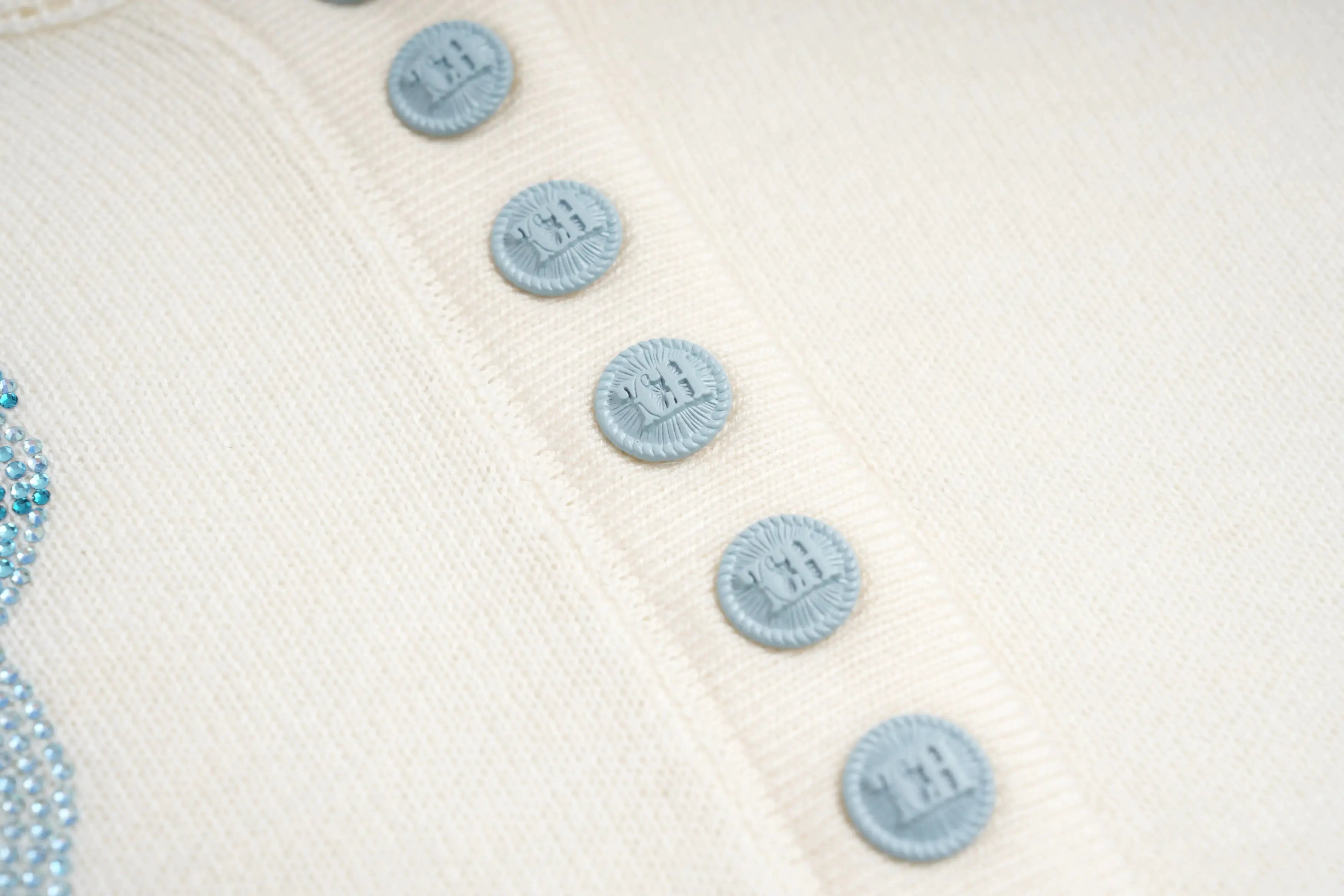 Close-up of a white textured fabric with blue buttons and a blue thread. TCH Rhinestone Teddy Bear Hooded Knit Tee