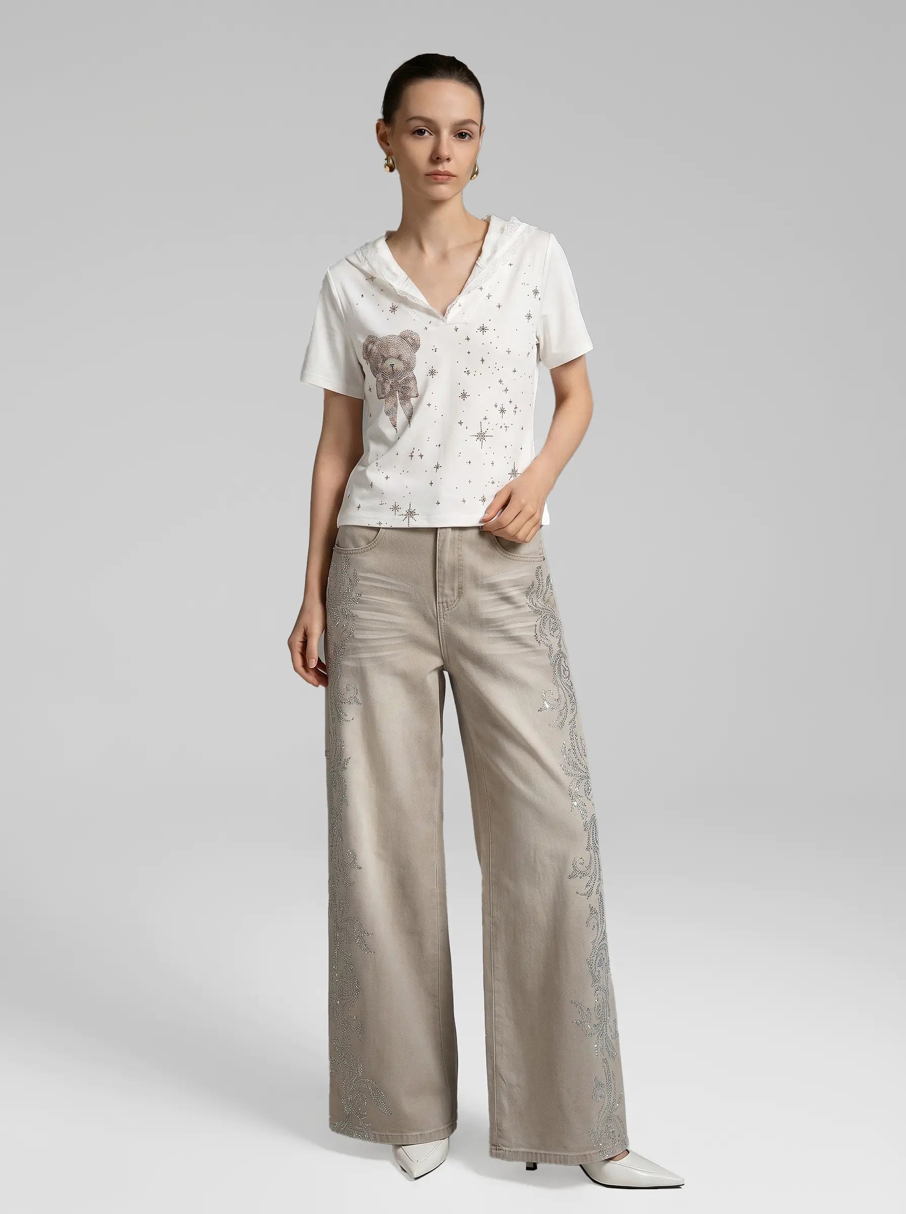 A model stands against a light gray studio background wearing a white short-sleeve top with a teddy bear motif and scattered star-like embellishments, paired with wide-leg beige trousers featuring tonal embroidery down the side, and white pointed-toe heels. TCH Rhinestone Teddy Bear Lace Hooded T-Shirt