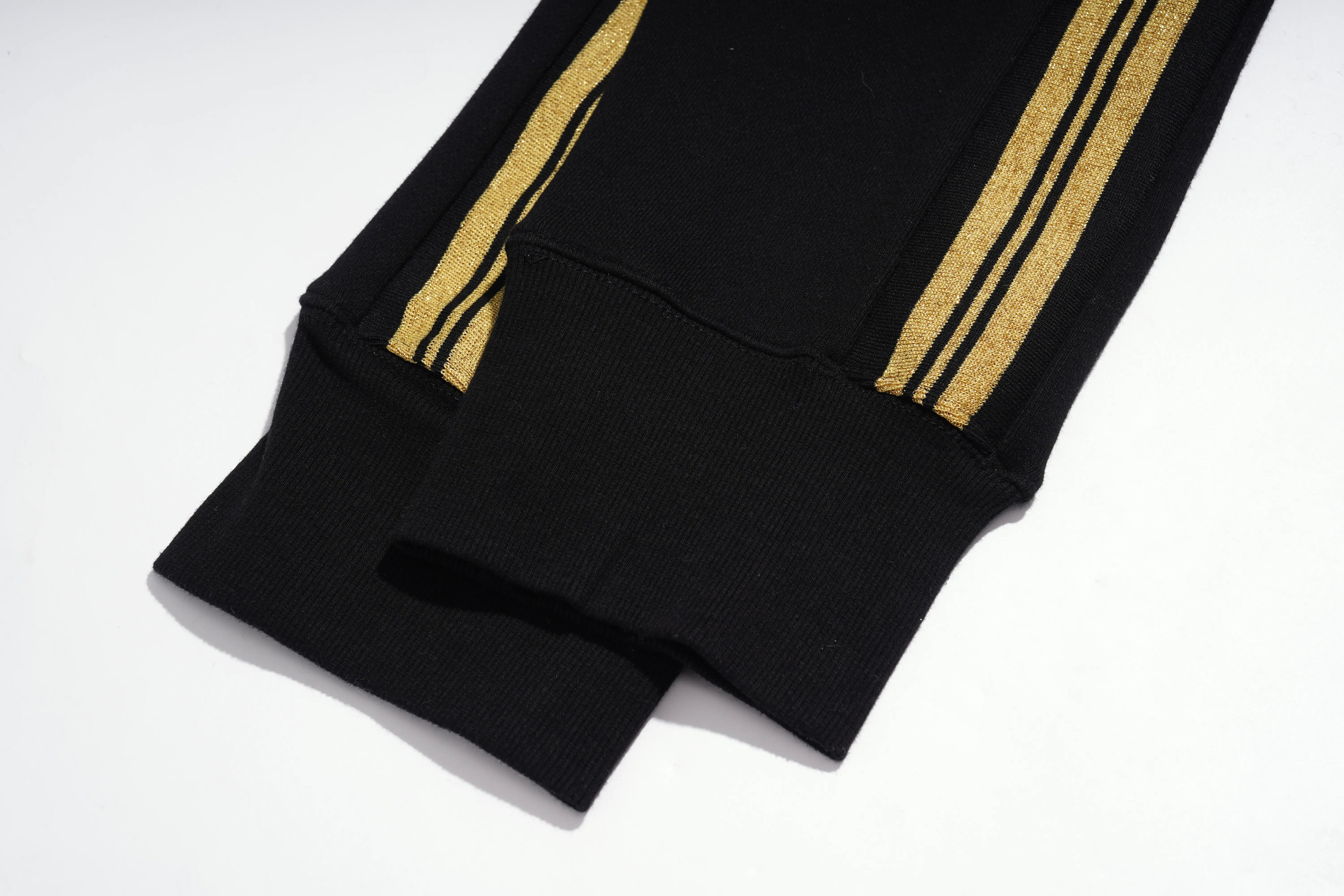 Close-up of a black ribbed knit garment, likely a sweatshirt or jacket sleeve, featuring two parallel gold stripes running down the side. TCH Rhinestone Teddy Bear London Graphic Black Jogger Pants