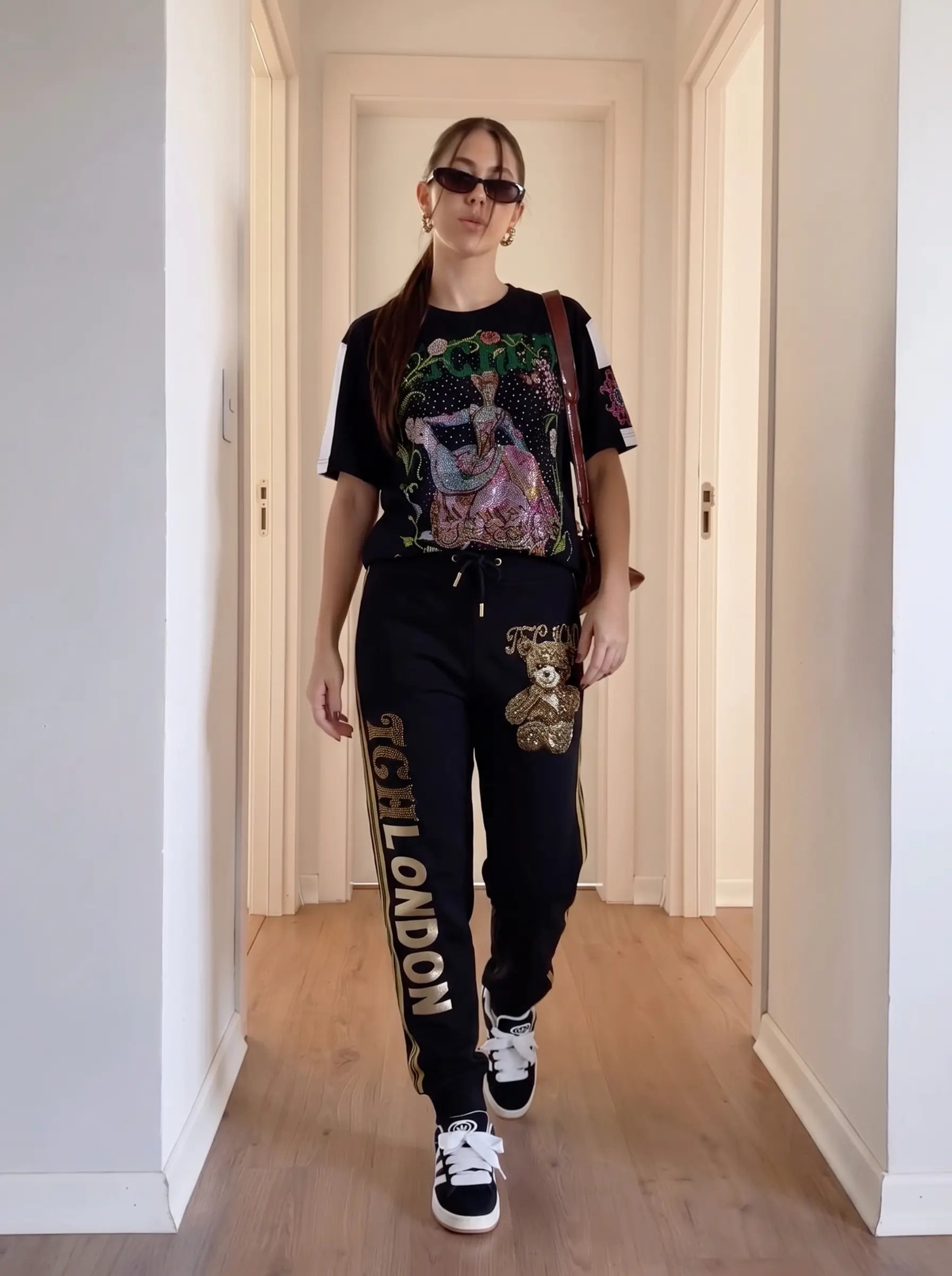 A woman walks down a hallway wearing a black graphic t-shirt, black sweatpants with 'CFL LONDON' text and a teddy bear patch, black sneakers, and sunglasses. TCH Rhinestone Teddy Bear London Graphic Black Jogger Pants