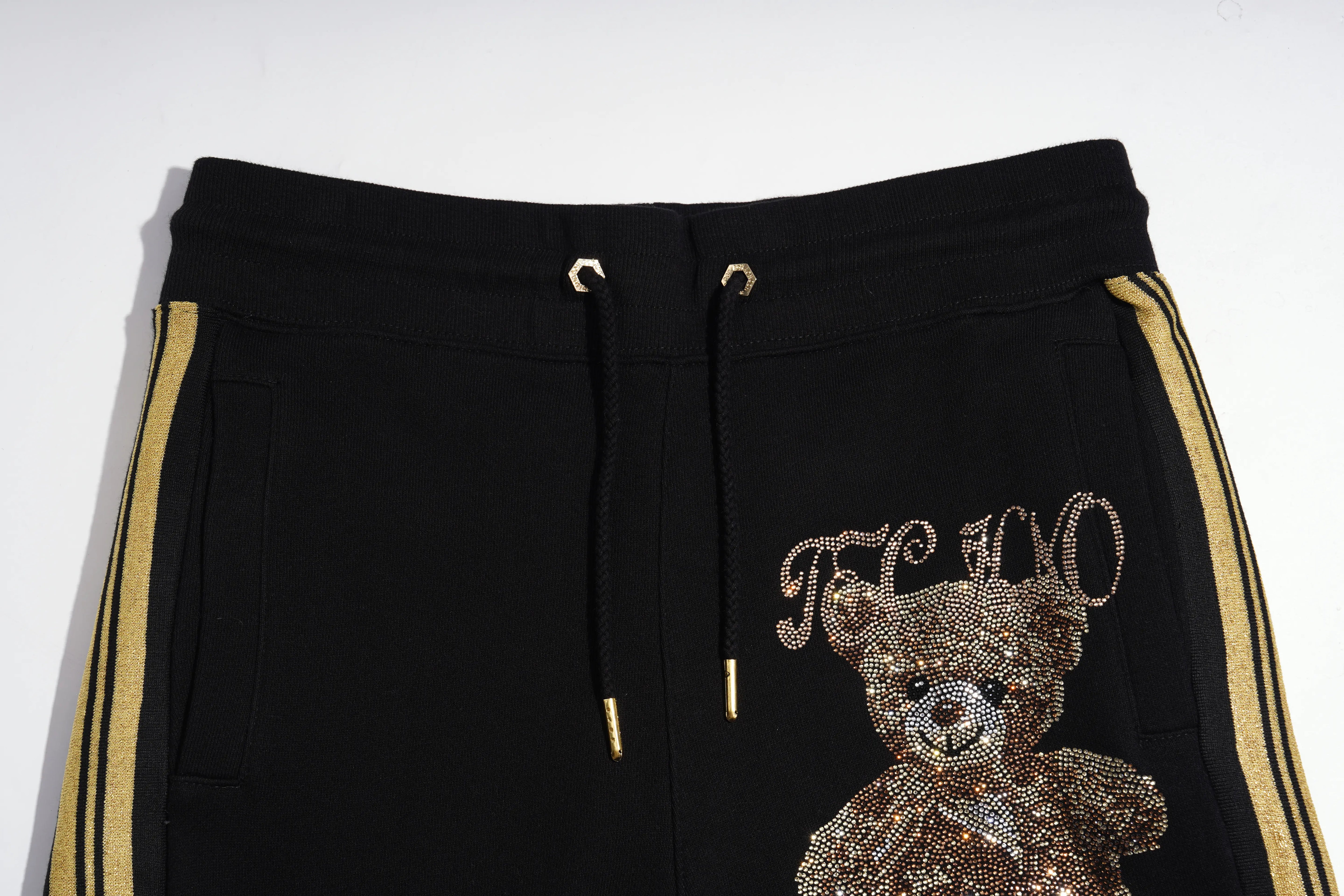Black sweatpants featuring a large sequined teddy bear on the left leg and gold lettering spelling 'FANCY' above it, with gold side stripes and drawstring rings. TCH Rhinestone Teddy Bear London Graphic Black Jogger Pants