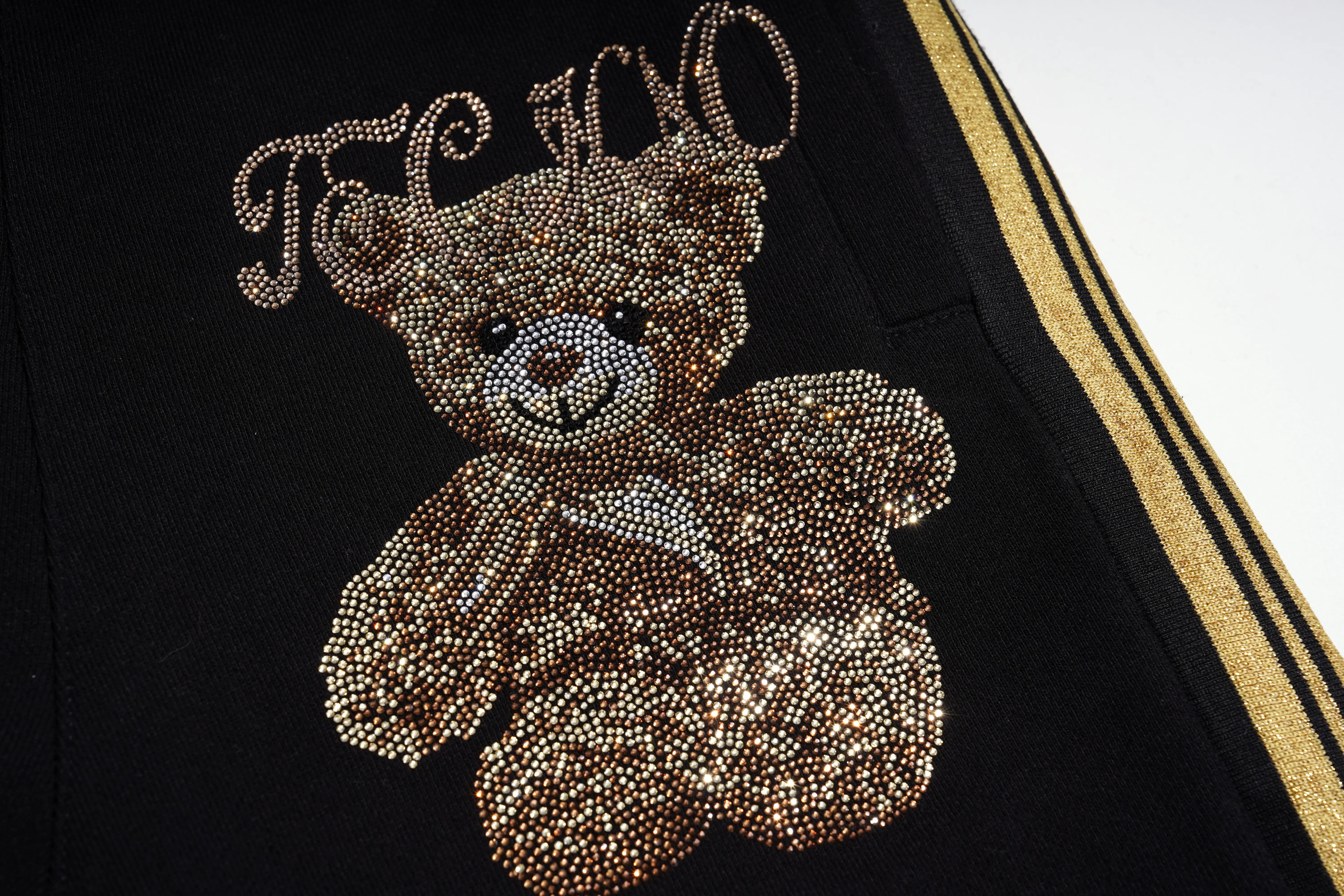 Close-up of a black garment featuring a large teddy bear embellished with gold, silver, and beige rhinestones, with the word 'FANCY' in matching rhinestones above it and a gold stripe detail on the side. TCH Rhinestone Teddy Bear London Graphic Black Jogger Pants