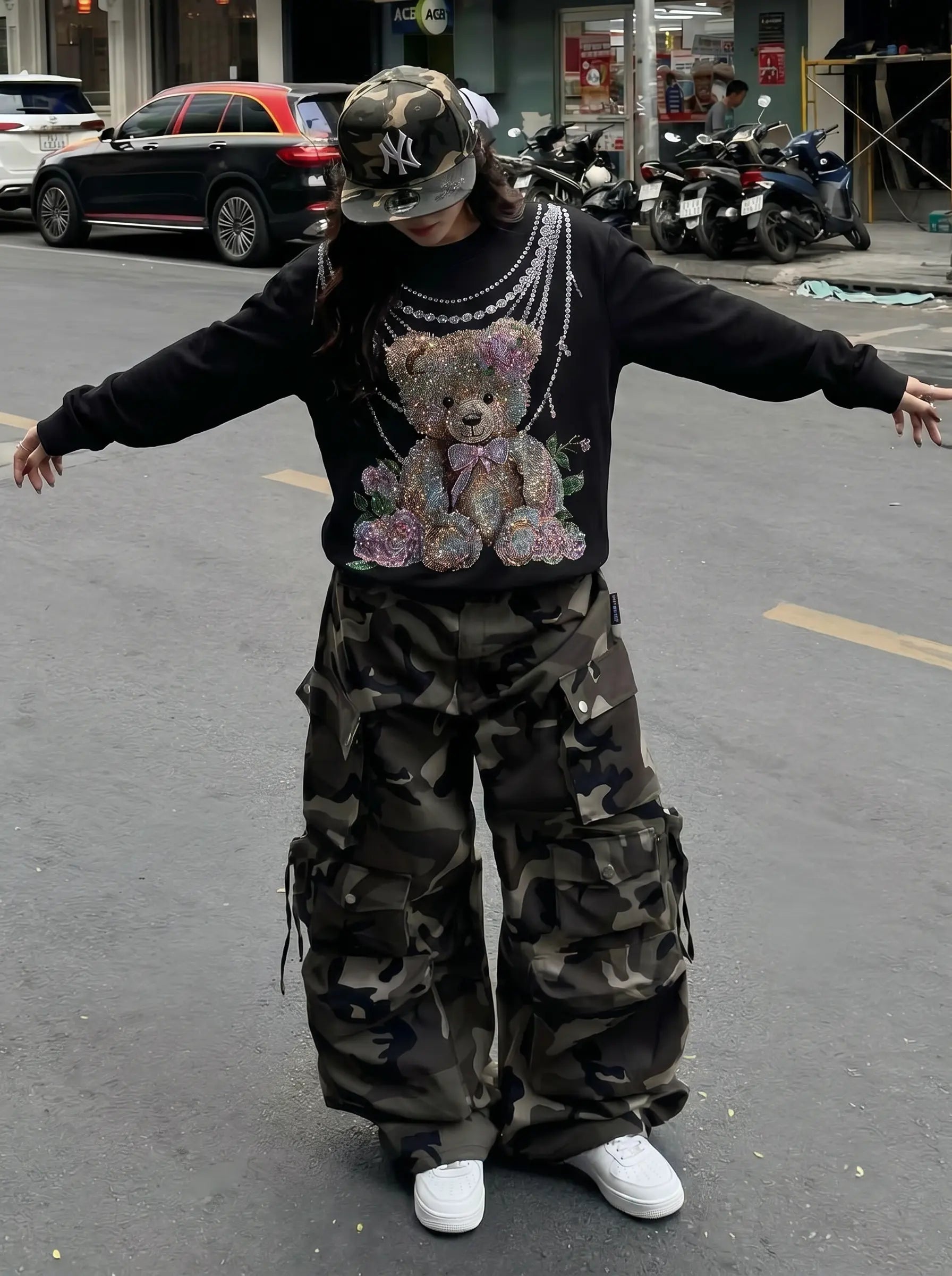 A person standing on a city street wearing a black long-sleeve sweatshirt with a large teddy bear graphic, camouflage cargo pants, a camouflage cap, layered silver necklaces, and white sneakers. TCH Rhinestone Teddy Bear Pearl Chain Graphic Sweatshirt