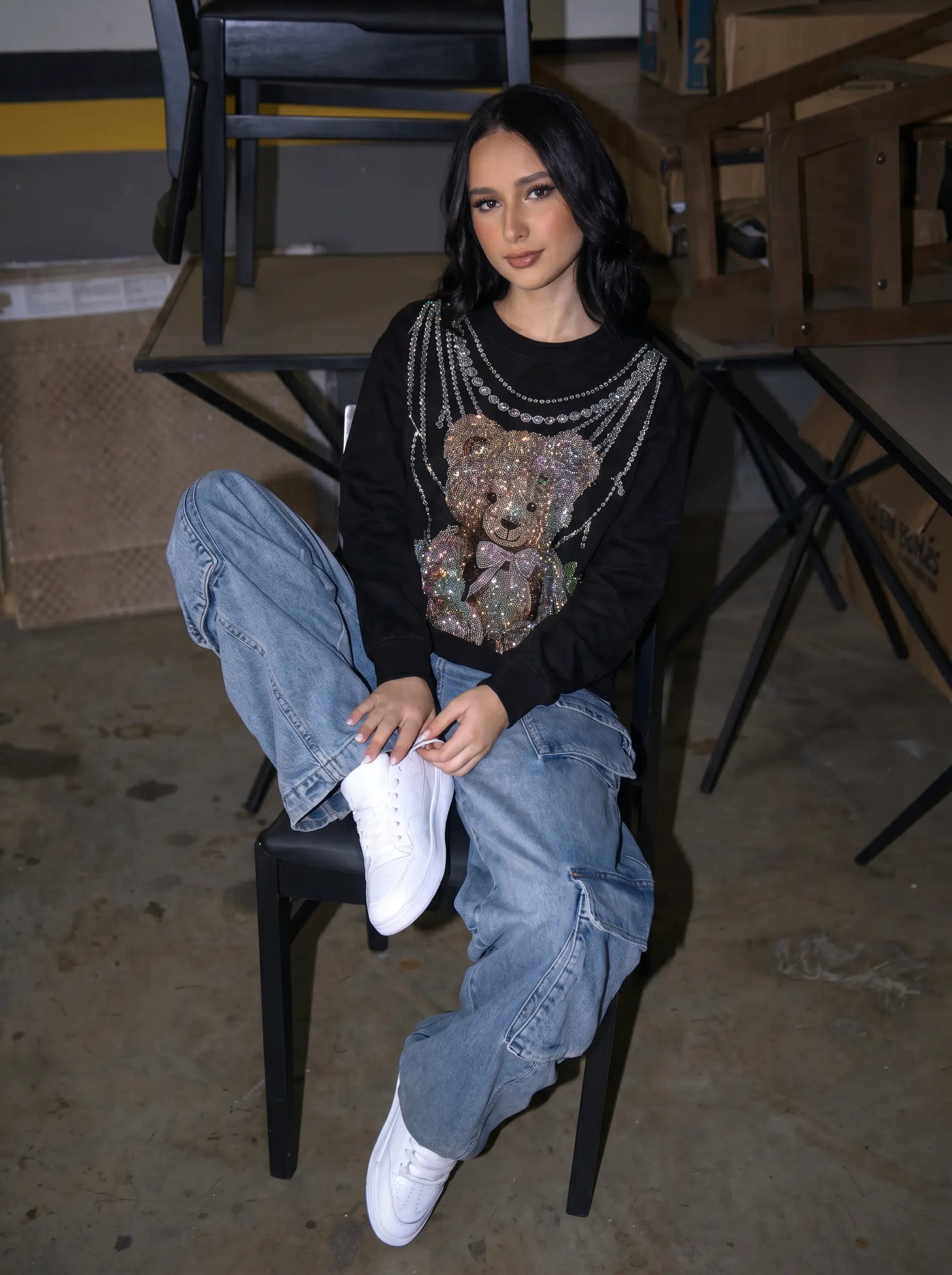 A woman sits on a black chair wearing a black long-sleeve top with a large teddy bear graphic, light blue baggy jeans, and white sneakers in an indoor setting with cardboard boxes and folding chairs. TCH Rhinestone Teddy Bear Pearl Chain Graphic Sweatshirt