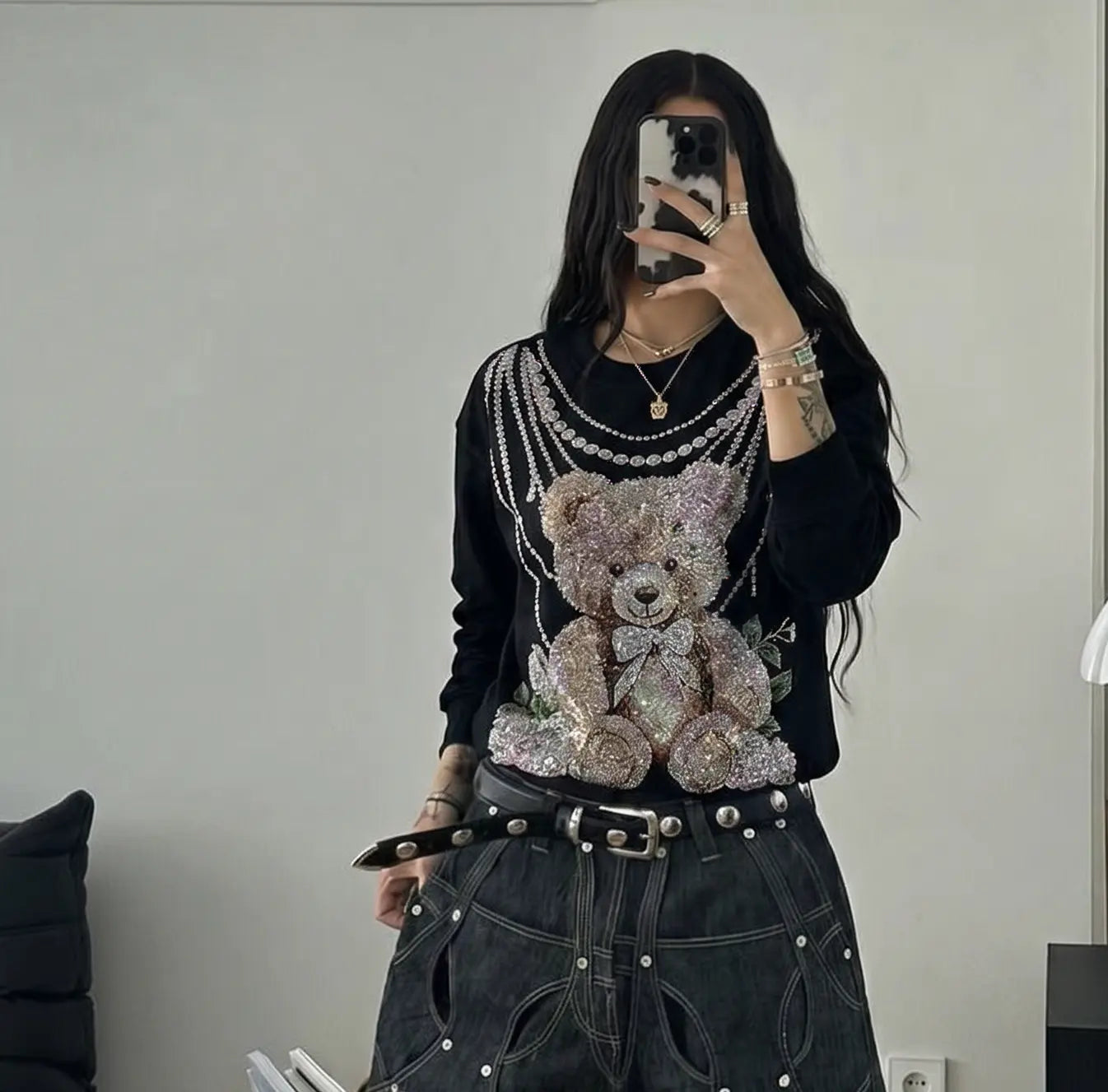 A person taking a mirror selfie wearing a black long-sleeve top with a large sequined teddy bear graphic and layered faux-pearl necklaces, paired with dark gray distressed jeans and a black belt. TCH Rhinestone Teddy Bear Pearl Chain Graphic Sweatshirt