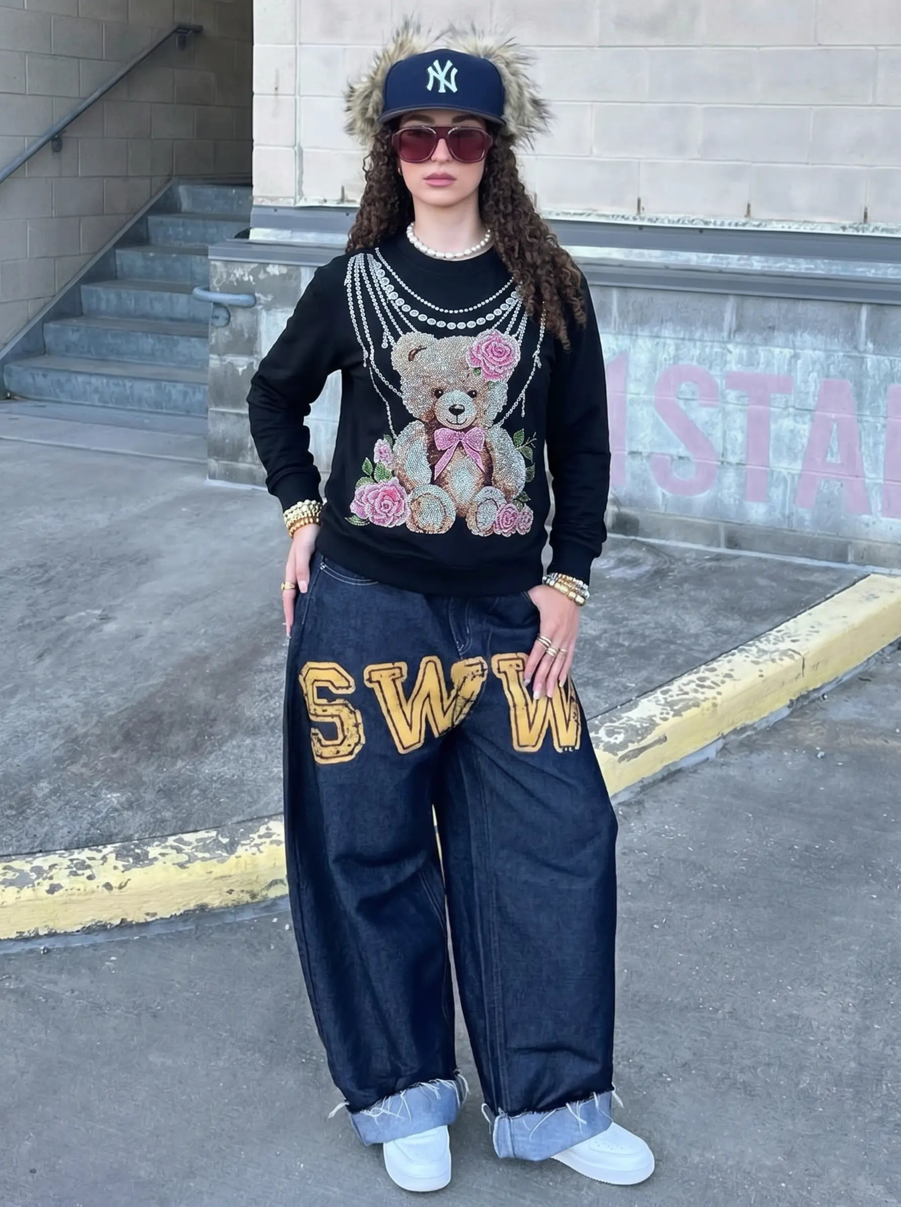 A person stands outdoors wearing a black long-sleeve sweatshirt with a teddy bear and pink rose graphic, dark wide-leg jeans with large gold 'SWO' lettering, a dark baseball cap with a fur trim and New York Yankees logo, sunglasses, and white sneakers. TCH Rhinestone Teddy Bear Pearl Chain Graphic Sweatshirt