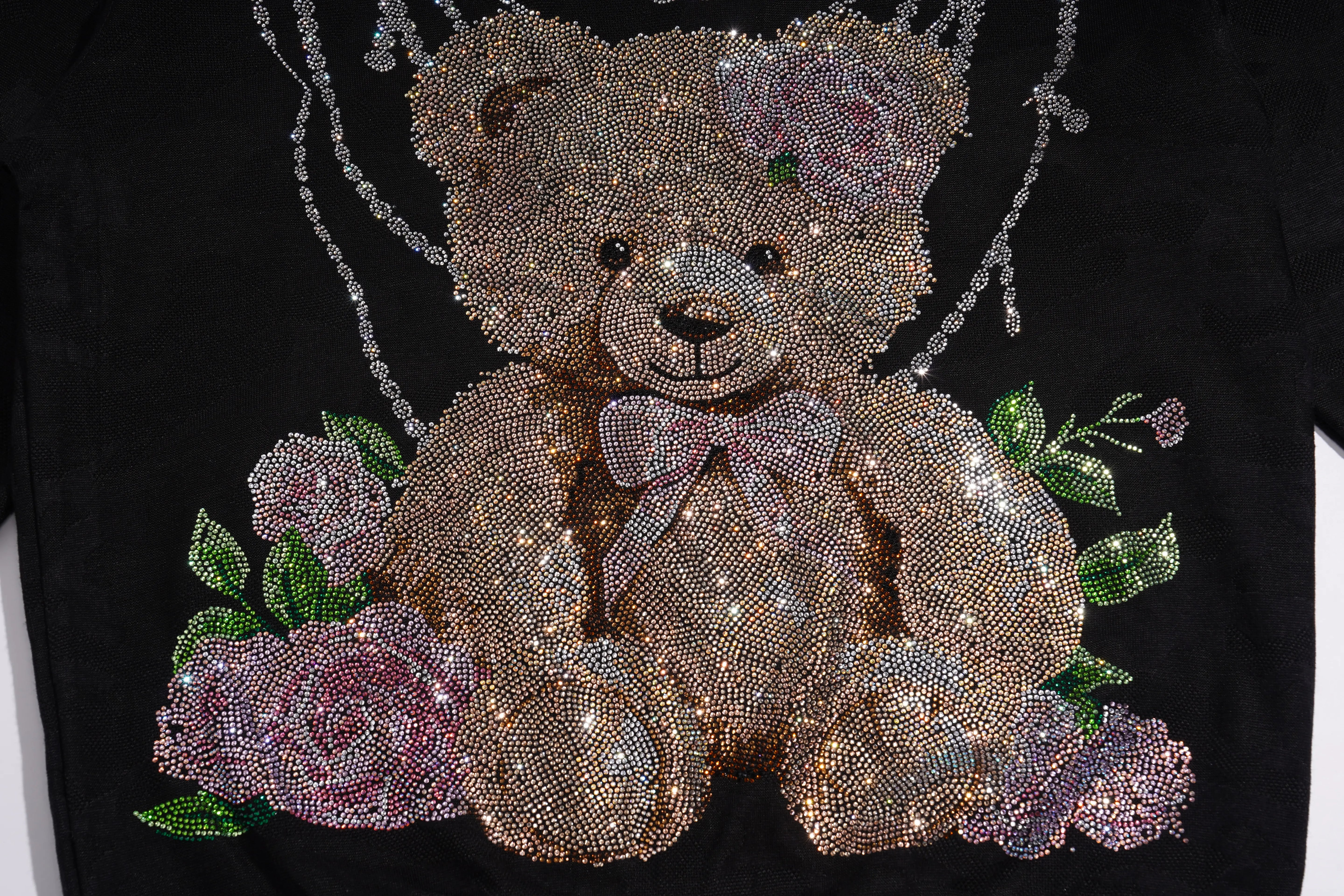 A close-up of a black garment featuring a large teddy bear graphic embellished with sparkling rhinestones, surrounded by pink roses and green leaves. TCH Rhinestone Teddy Bear Pearl Chain Graphic Sweatshirt