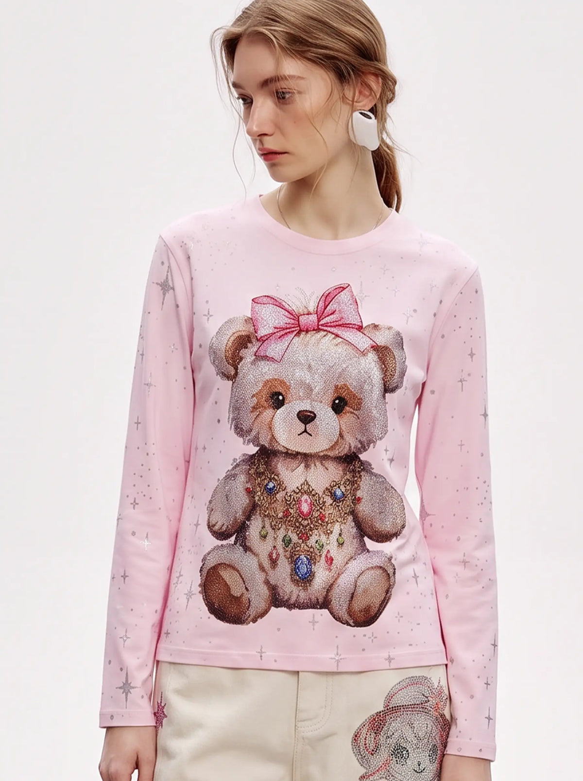 A model wearing a light blue denim jacket with various embroidered patches, including a teddy bear on the left chest and cherries on the sleeve, styled over a white t-shirt and dark pants. TCH Rhinestone Teddy Bear Pink Long Sleeve T-Shirt