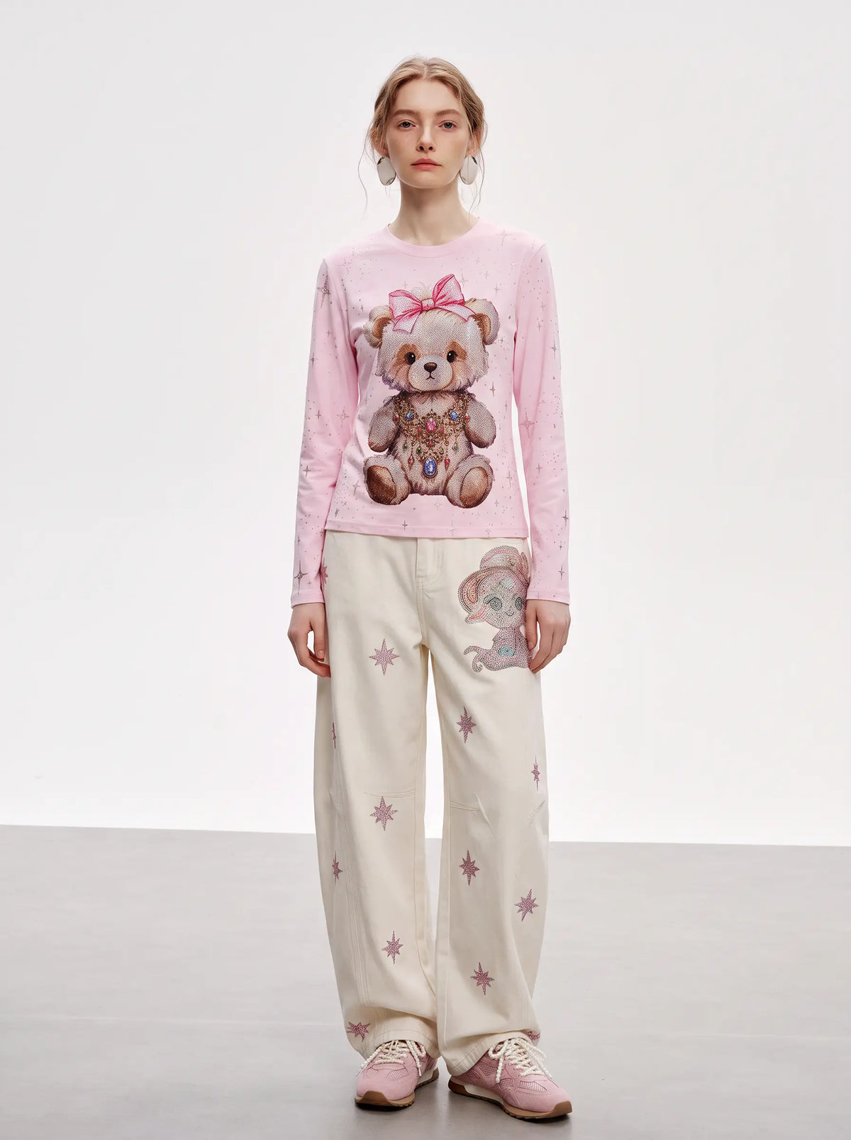 A model stands against a light gray studio background wearing a pink long-sleeve top with a teddy bear graphic and cream wide-leg pants with pink star prints and a matching teddy bear patch. TCH Rhinestone Teddy Bear Pink Long Sleeve T-Shirt