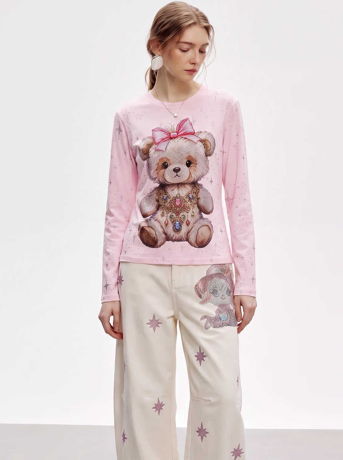 A model wearing a light pink long-sleeve top with a large teddy bear graphic on the front, paired with cream-colored pants featuring star prints and a matching teddy bear print on the thigh. TCH Rhinestone Teddy Bear Pink Long Sleeve T-Shirt