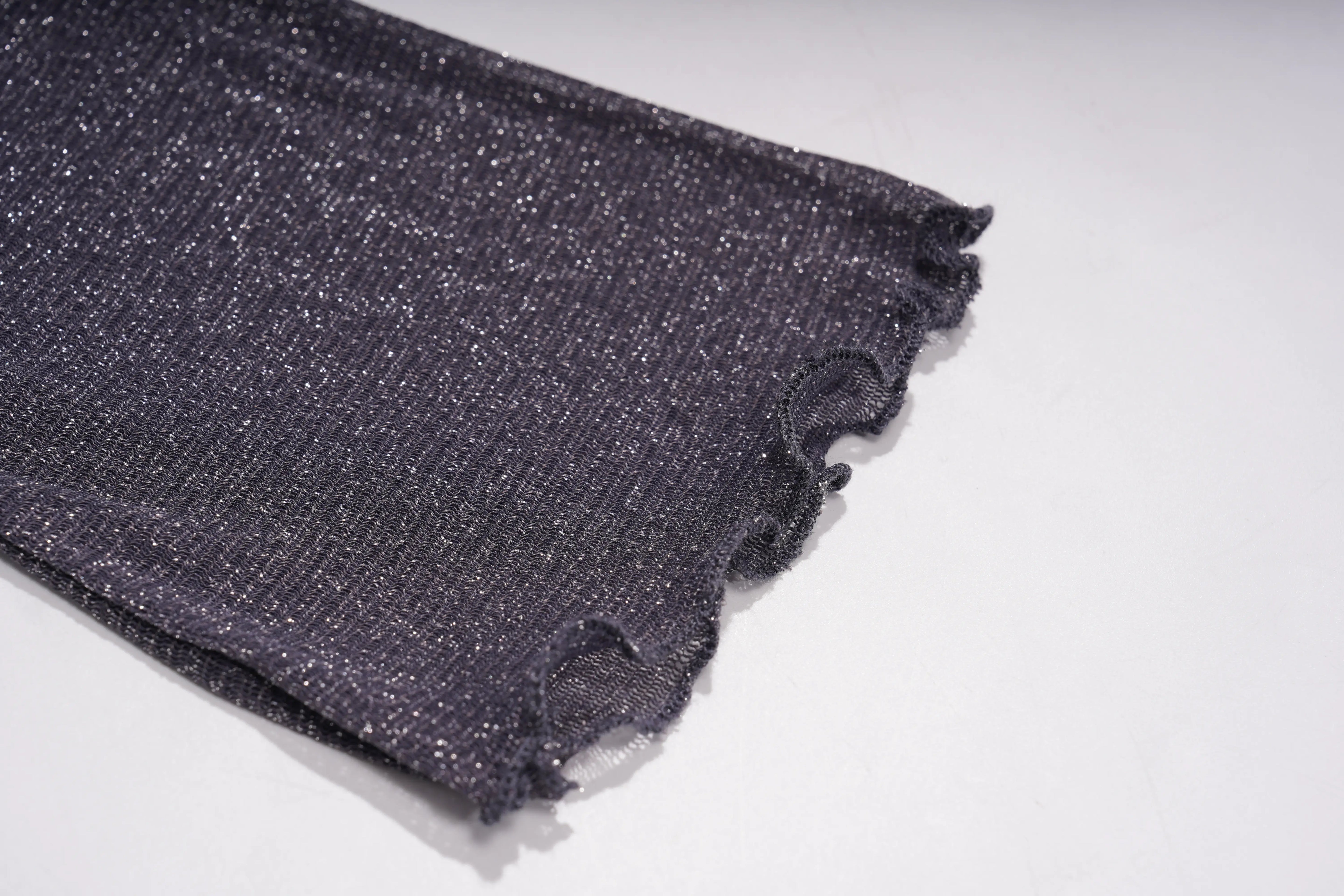 A close-up of a dark charcoal gray or black knit fabric with a subtle glittery texture and frayed, ruffled edges, laid on a plain white surface. TCH Rhinestone Teddy Bear Sheer Mesh Long Sleeve Top