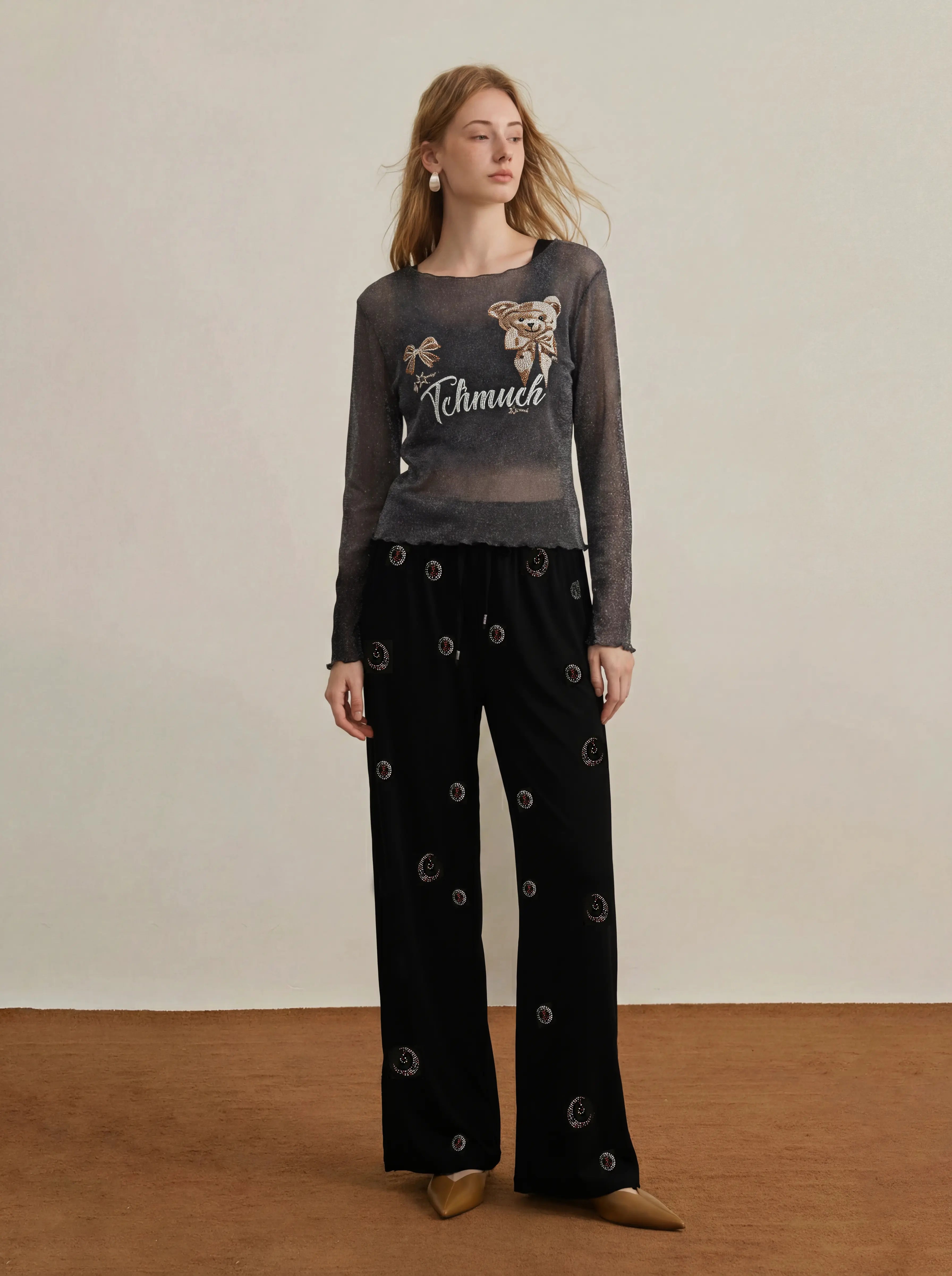 A woman stands in a studio wearing a sheer dark gray long-sleeve top with a graphic print and black wide-leg pants with circular cutout details. The background is a plain beige wall with a brown floor. TCH Rhinestone Teddy Bear Sheer Mesh Long Sleeve Top