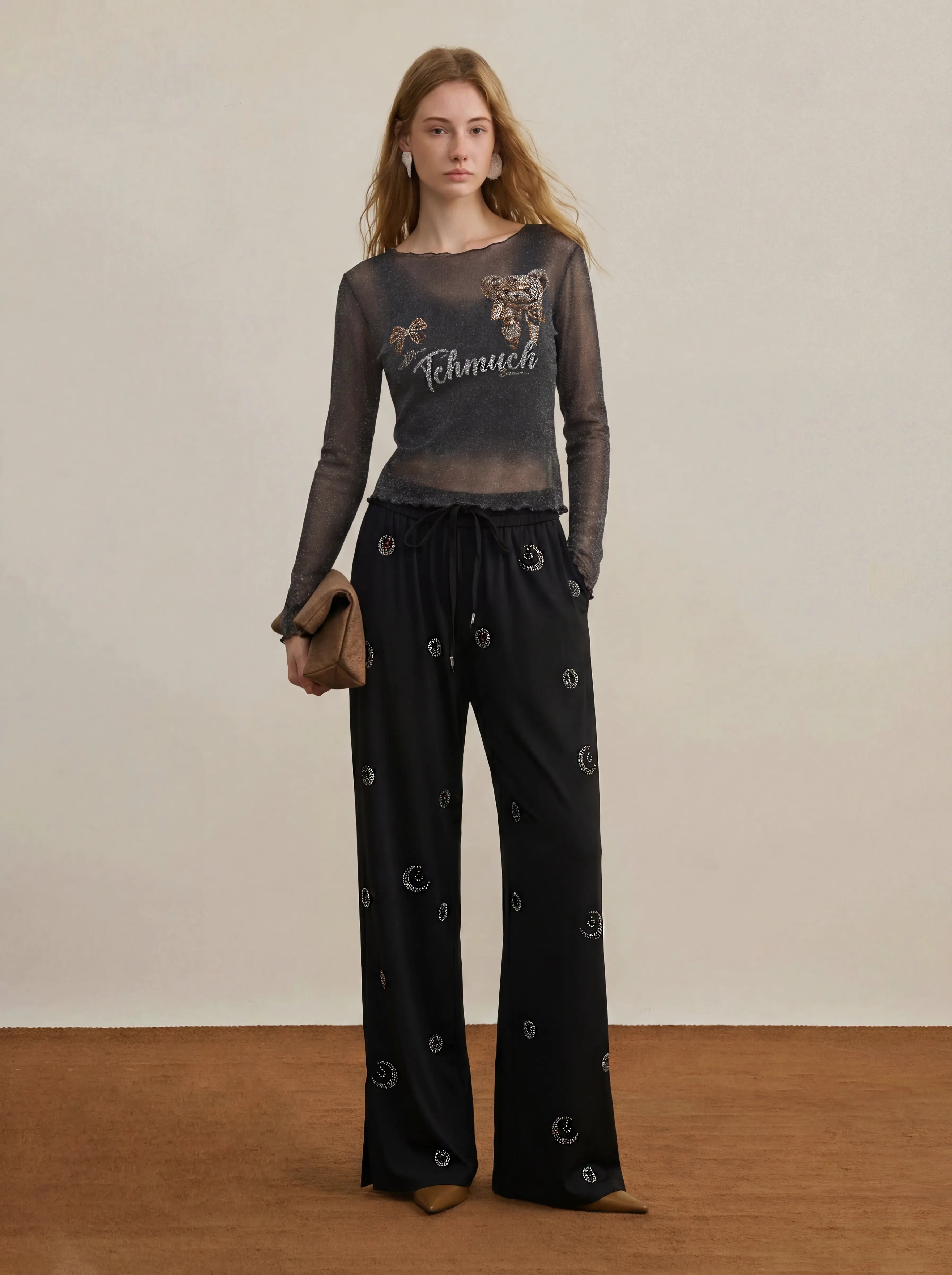 A model stands in a studio wearing a sheer dark gray long-sleeve top with a graphic print and black wide-leg pants with small circular prints, holding a brown clutch against a neutral beige background. TCH Rhinestone Teddy Bear Sheer Mesh Long Sleeve Top