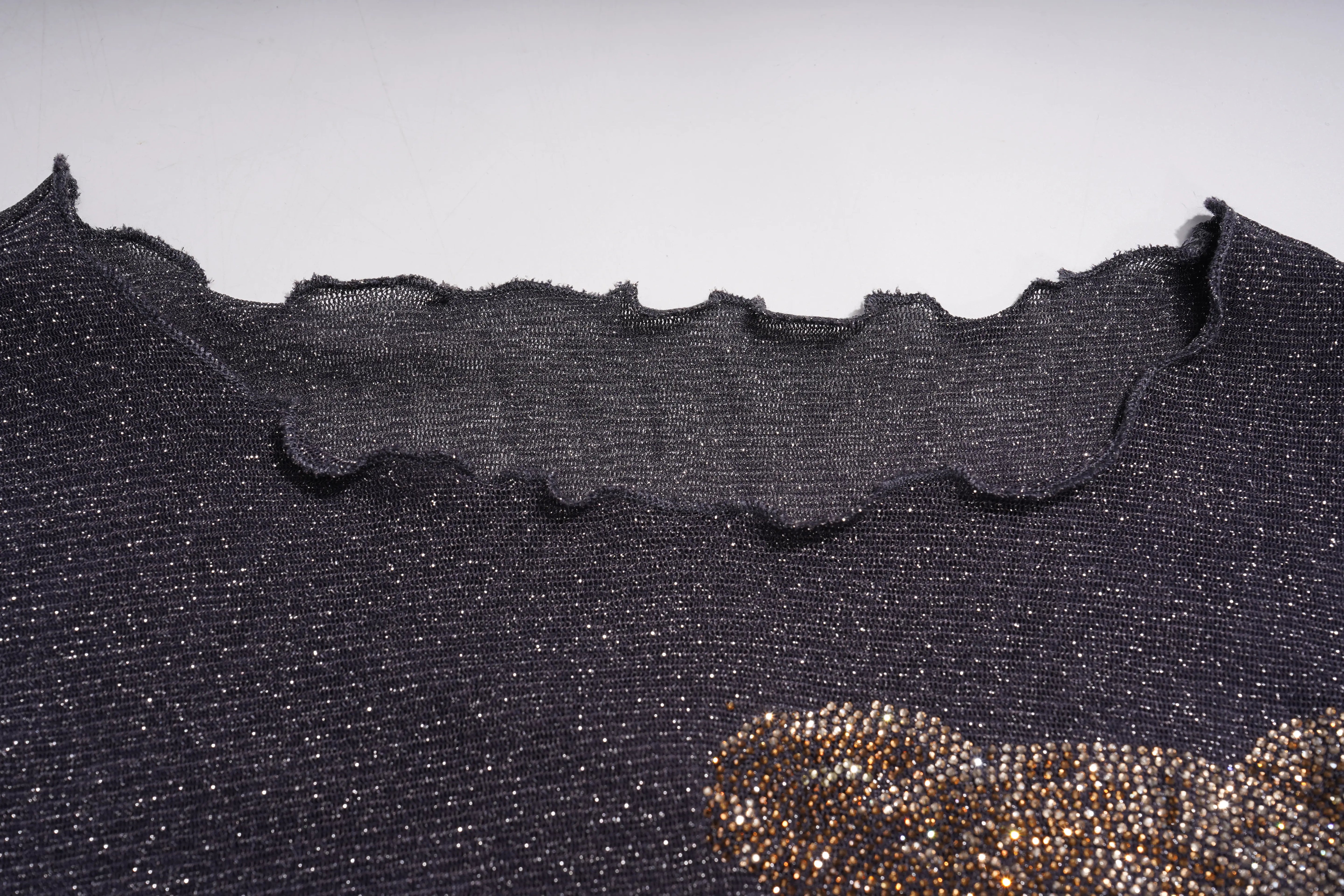 Close-up of a dark textured knit garment with a scalloped wavy neckline, featuring a gold beaded embellishment in the lower right corner against a light gray background. TCH Rhinestone Teddy Bear Sheer Mesh Long Sleeve Top