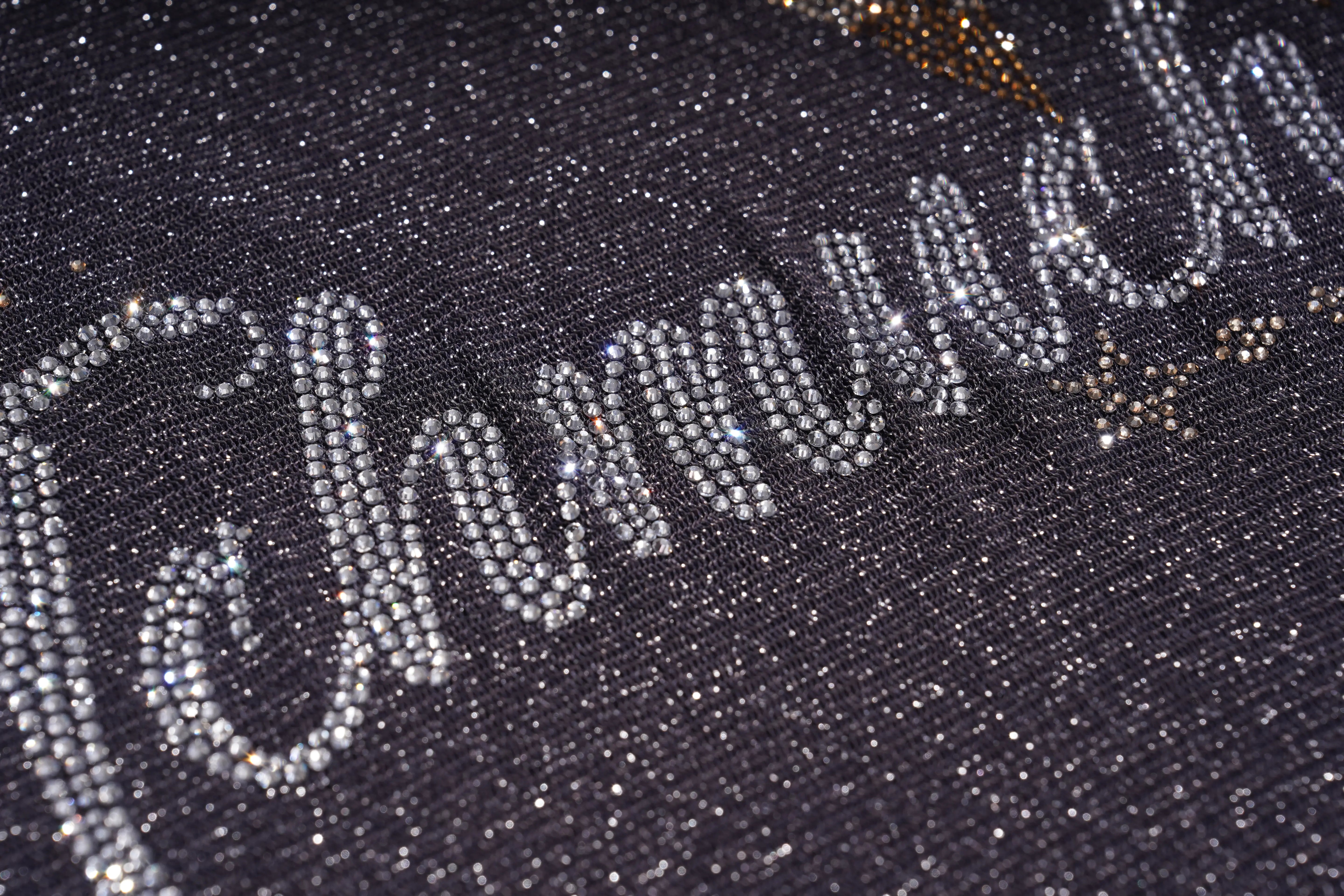 A close-up of a dark glittery fabric with large silver rhinestone script lettering and small gold star accents. TCH Rhinestone Teddy Bear Sheer Mesh Long Sleeve Top