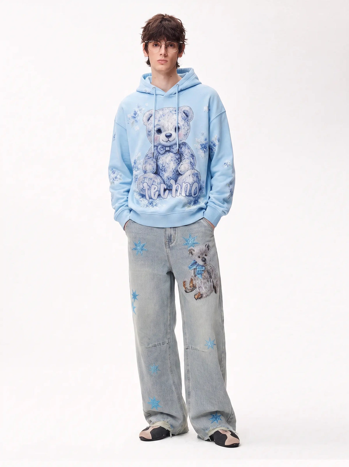A model stands against a plain white studio background wearing a light blue hoodie with a large teddy bear graphic and the word 'MATHWO' on the front, paired with light-wash jeans featuring blue star prints and a small bear patch, and dark slip-on shoes. TCH Rhinestone Teddy Bear Star Wide-Leg Jeans
