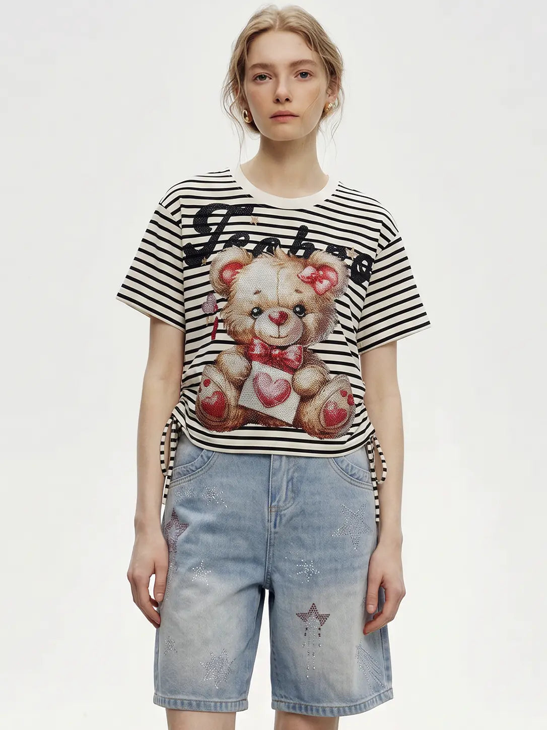 A model wearing a black and white striped t-shirt with a teddy bear graphic and denim shorts, photographed against a plain light gray background. TCH Rhinestone Teddy Bear Striped Tie-Side T-Shirt