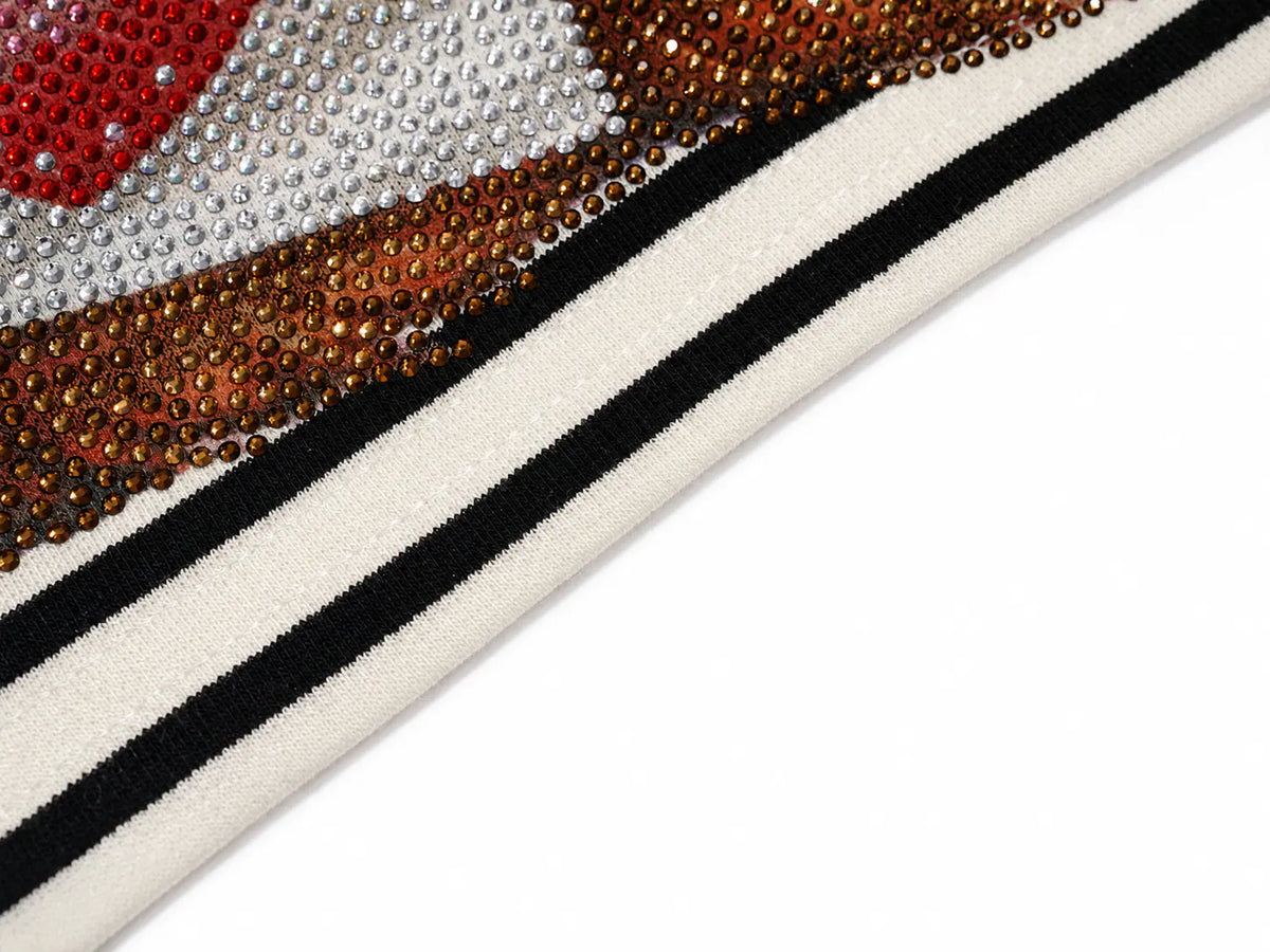 Close-up of a textile or trim sample with a cream base, two black stripes, and a decorative border made of rhinestones in red, silver, and metallic bronze tones, shown on a white background. TCH Rhinestone Teddy Bear Striped Tie-Side T-Shirt