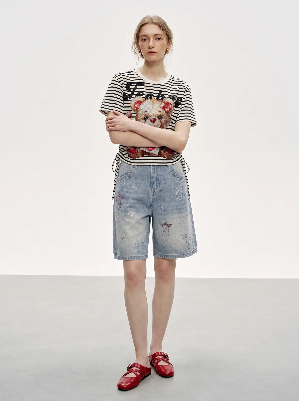 A model stands against a light gray studio background wearing a black and white striped graphic t-shirt with a teddy bear print, light-wash denim shorts, and red patent leather loafers. TCH Rhinestone Teddy Bear Striped Tie-Side T-Shirt