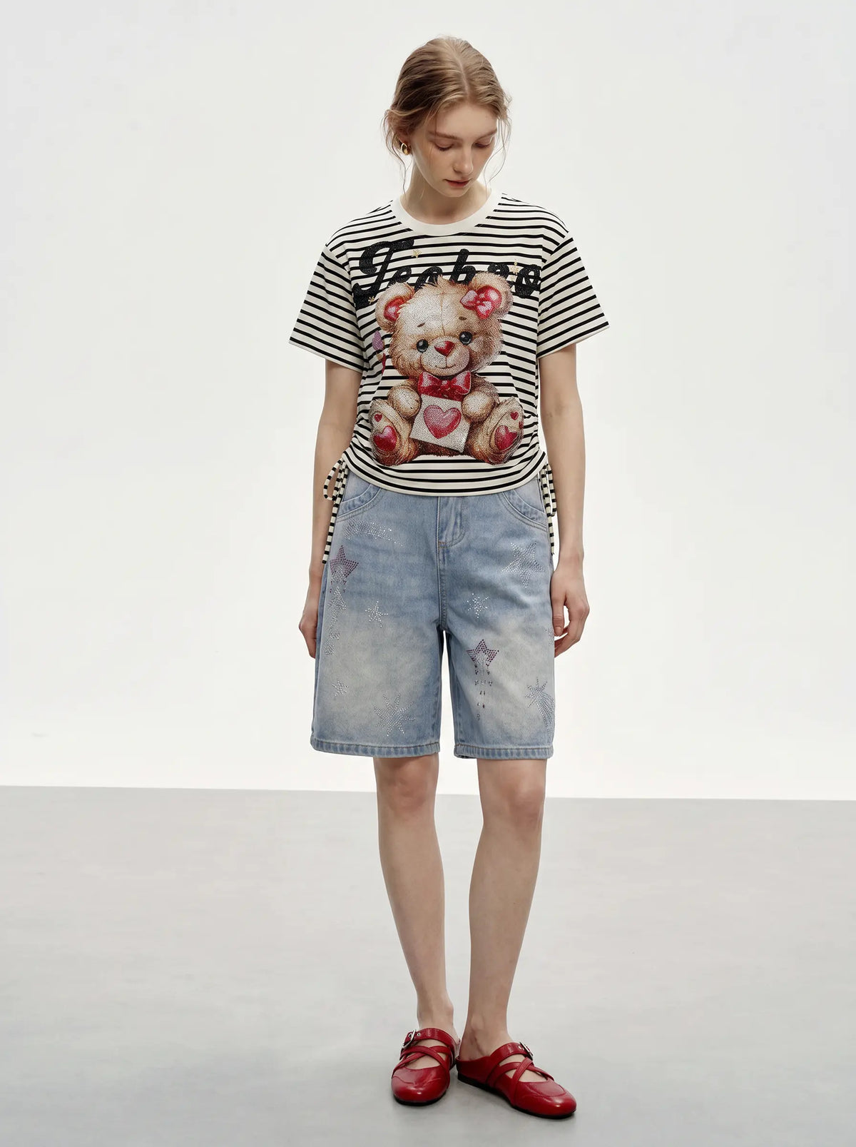 A model stands against a light gray studio background wearing a black and white striped t-shirt with a teddy bear graphic, light-wash denim shorts, and red loafers. TCH Rhinestone Teddy Bear Striped Tie-Side T-Shirt